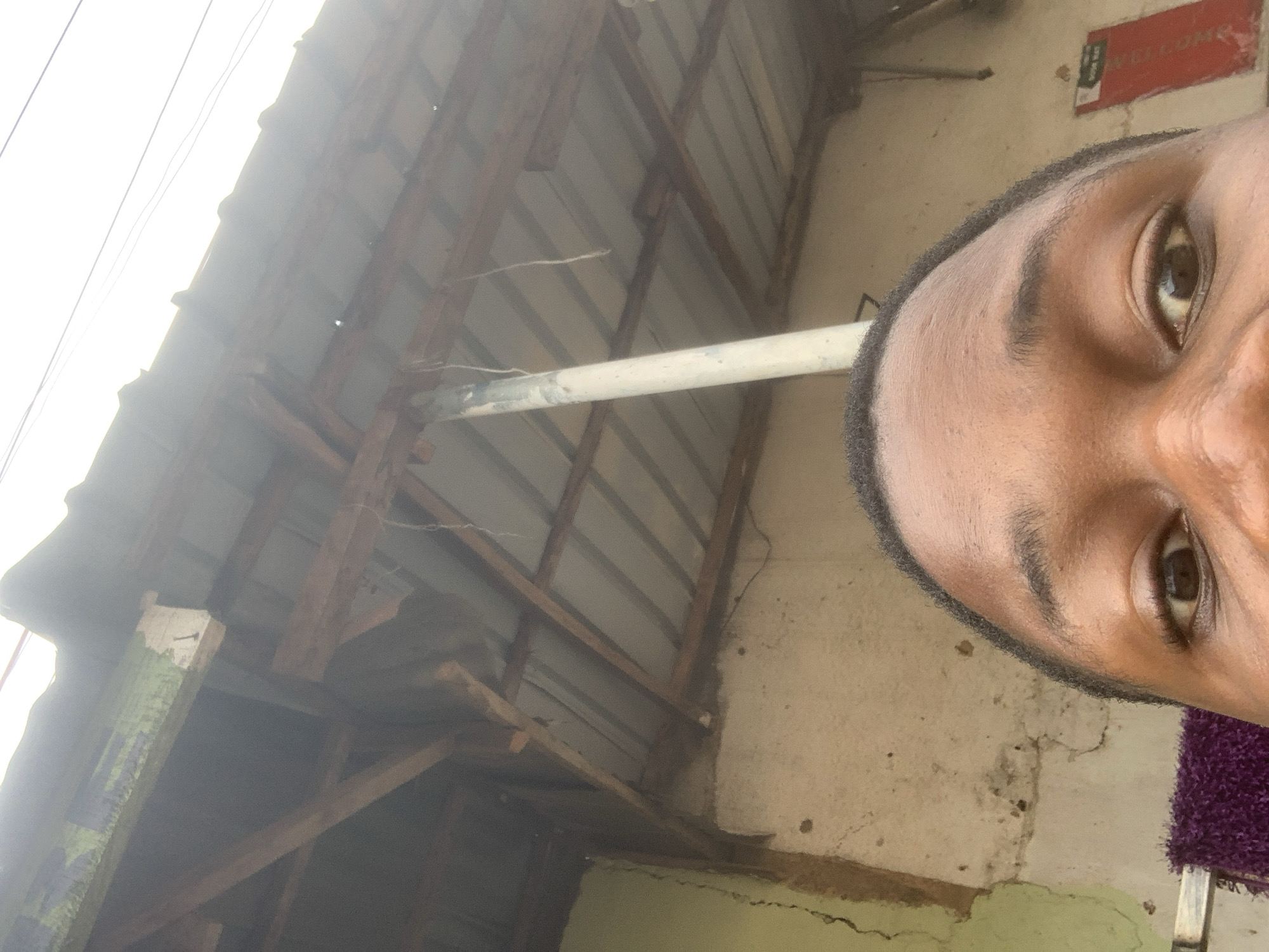 The image is a close-up, low-angle shot of a person's eyes and forehead, taken outdoors in what appears to be a rural or semi-urban environment in Gura Top, Nigeria. The background consists of a corrugated metal roof supported by wooden beams, with some visible cracks and discoloration on the building's wall. A white pipe runs vertically through the scene, and thin wires are visible against the bright sky. The overall lighting suggests it is daytime. The person's skin tone is dark, and their hair is short and dark. A purple textured object is partially visible in the lower left corner. A red sign with some text is on the wall in the right background. The composition is informal, suggesting a selfie or a candid shot.