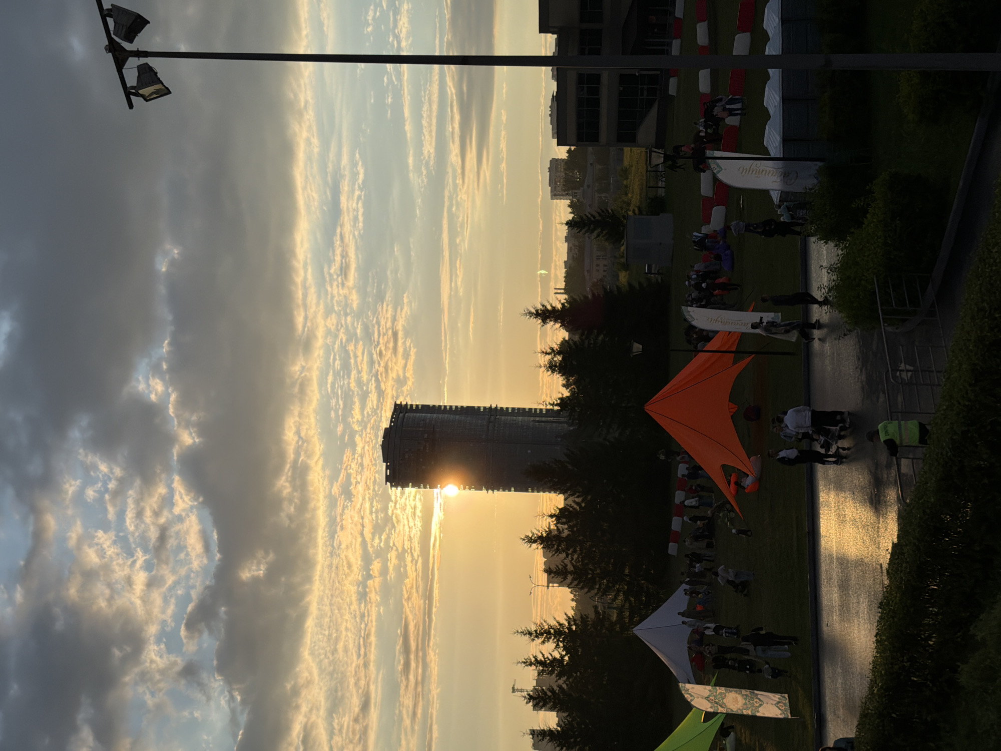 An outdoor scene at dusk shows a cluster of tents and people gathered on a grassy field in front of a tall, cylindrical skyscraper. The sun is setting behind the skyscraper, casting a bright glow and peeking through the building's windows. Various tents, including an orange pyramid-shaped one and a white conical one, are set up on the field. People are walking around, some with strollers, and others are congregating near the tents. In the foreground, a wet asphalt surface reflects the setting sun. A lamppost with two lights stands tall on the right side of the image. Pine trees and other buildings are visible in the background. The sky is filled with dramatic clouds, illuminated by the sunset.