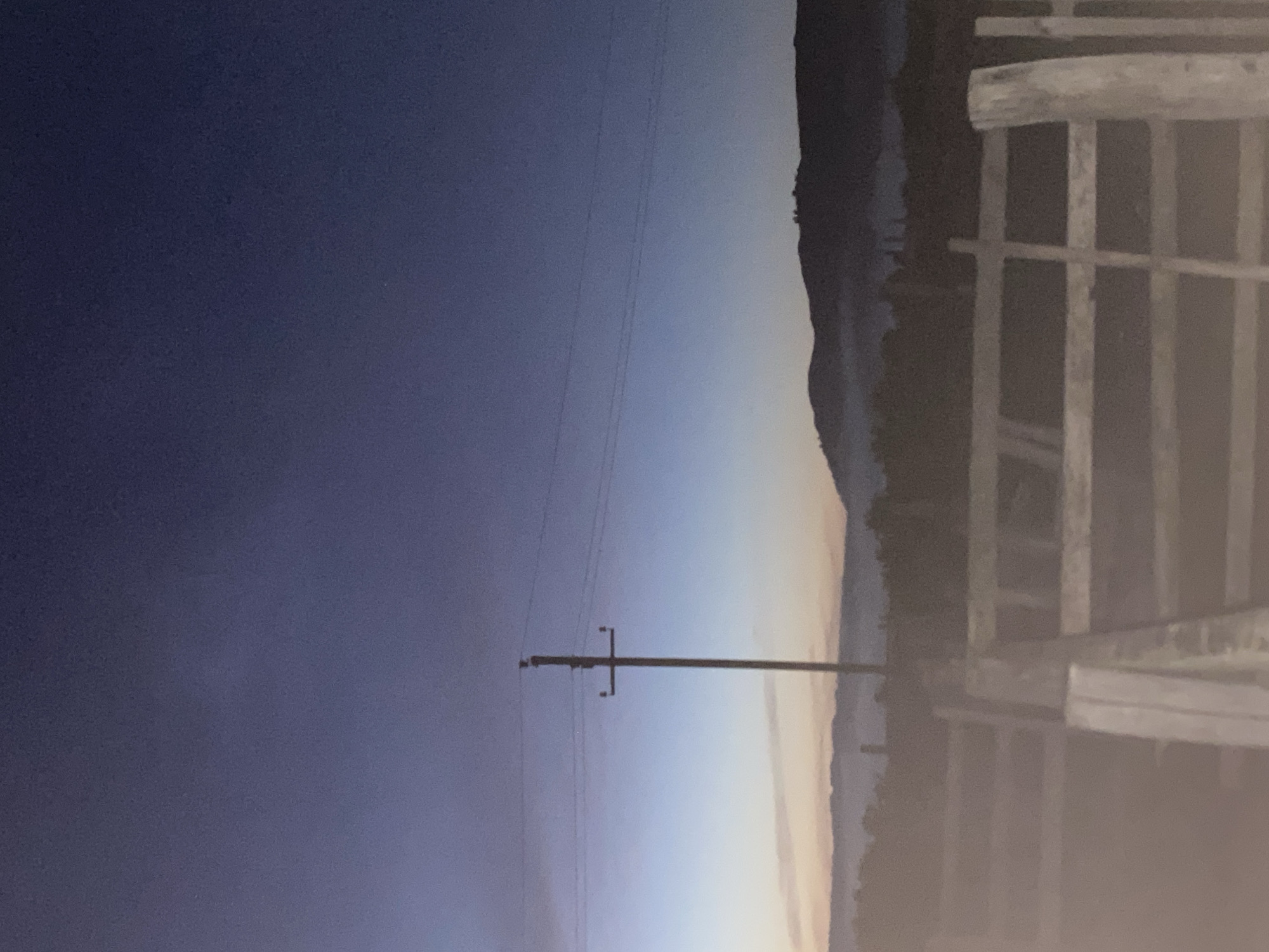 This image depicts a rural landscape at dusk or dawn, featuring a wooden fence in the foreground. Behind the fence, a utility pole with electrical wires stands prominently. In the background, hills are visible, partially obscured by a light mist or fog. The sky transitions from a deep blue at the top to lighter shades of blue and orange near the horizon. The overall impression is one of tranquility and natural beauty. This scene is located in Maloarslangulovo, Russia.