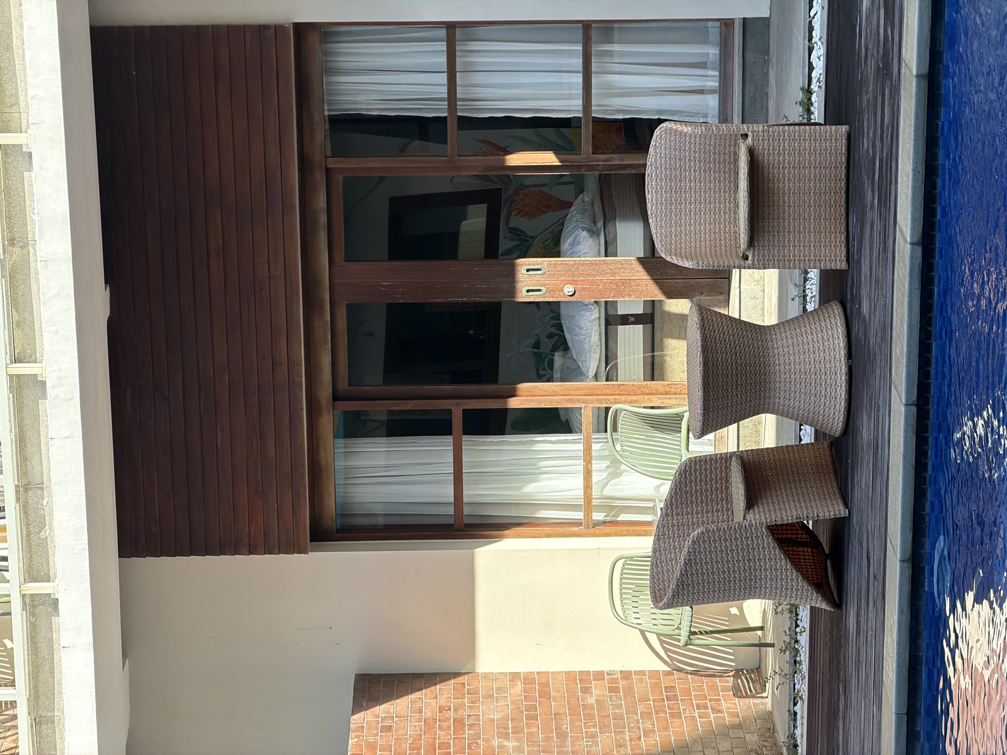 This is an eye-level, medium shot of a patio area next to a swimming pool, in front of a building with a sliding glass door. The patio is constructed of dark wood planks. A brown wicker armchair is situated on the left, with a matching hourglass-shaped side table in the center, and another brown wicker armchair on the right. Two light green metal chairs are visible behind the wicker furniture, set against a light-colored wall. A brick wall is to the left of the building. The building itself has a dark wood-paneled section above the glass door, and white curtains are drawn behind the glass. Sunlight casts shadows across the scene. This scene is likely located in Kuta Selatan, Indonesia, given the tropical setting and architectural style.