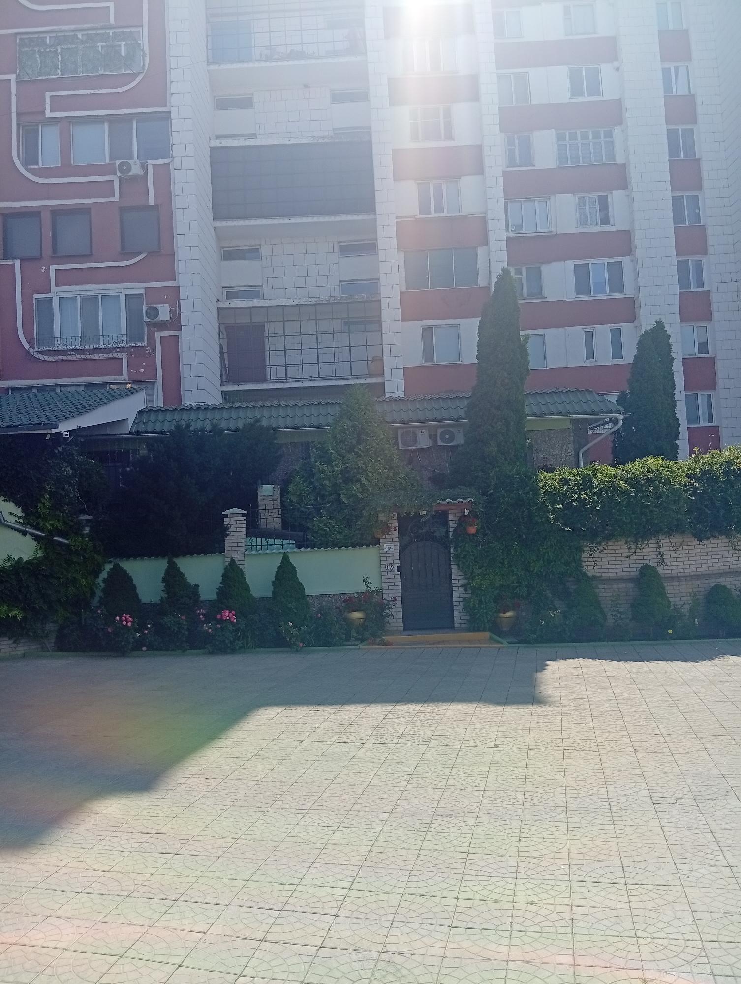 The image shows a multi-story building with a pink and white facade, likely an apartment complex, in Rybnitsa, Moldova. The building features numerous windows and balconies. In front of the building, there is a paved courtyard with a tiled surface. A green fence separates the courtyard from a garden area with various plants, including roses and ornamental trees, and a dark-colored gate. The scene is illuminated by sunlight, casting shadows on the courtyard. The overall impression is of a residential area on a clear day.