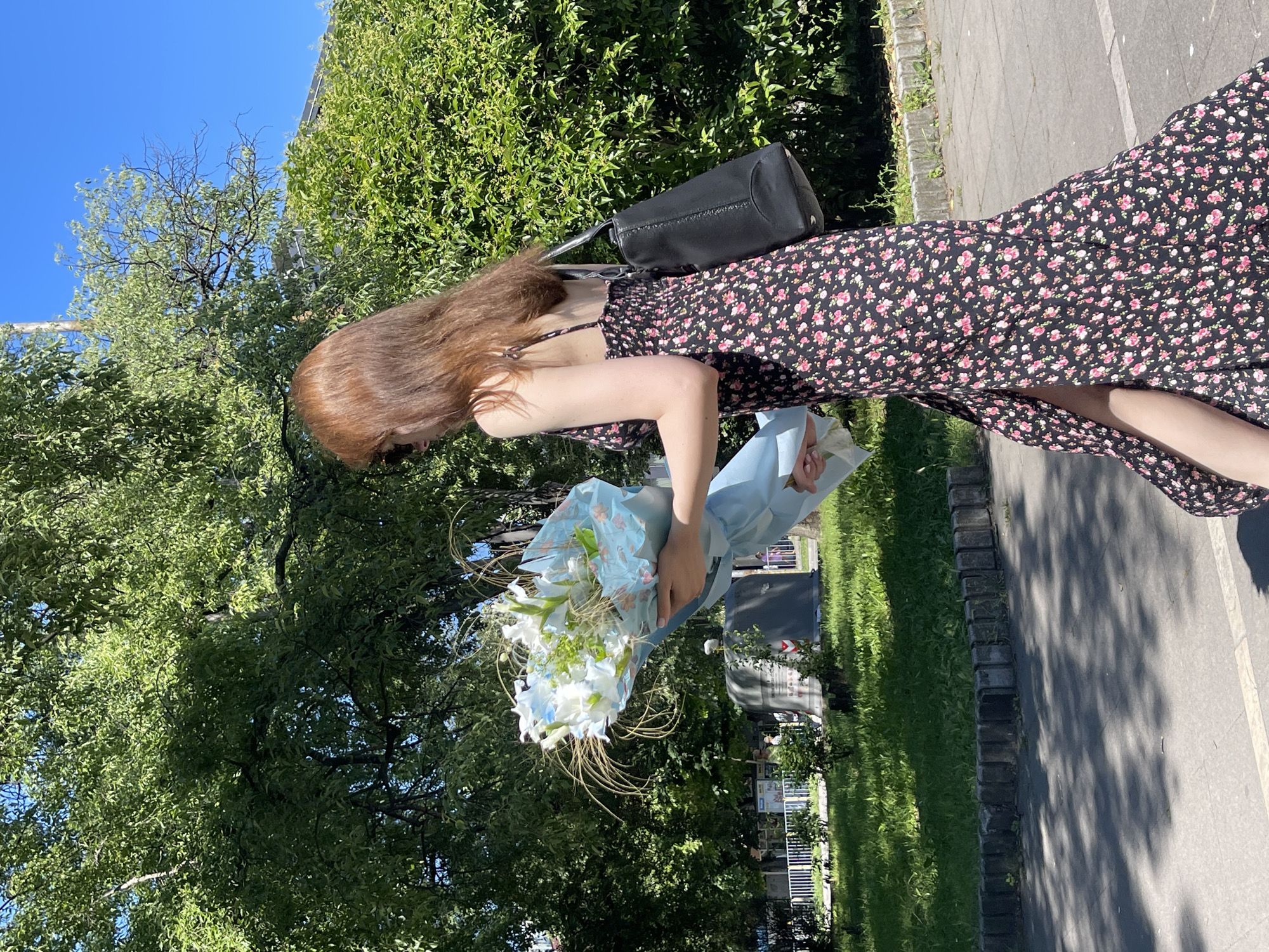 In the city of Varna, Bulgaria, on a sunny day, a young woman with long, reddish-brown hair walks along a paved path. She is wearing a black floral print maxi dress with a high slit on the side and a black shoulder bag. In her arms, she carries a large bouquet of white flowers wrapped in light blue paper. She is walking from left to right in the frame, facing slightly away from the camera. The background features a lush green park with trees and bushes, and a clear blue sky above. The sunlight casts shadows on the path, indicating it is midday.