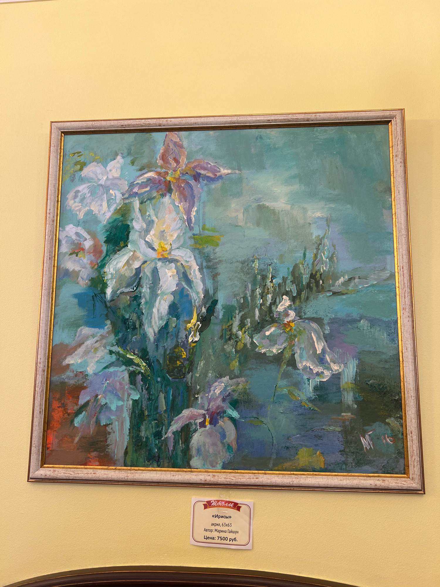 The image displays a framed oil painting of irises against a teal-blue background. The painting, signed "M.G." on the right side, is displayed on a pale yellow wall. A small white label below the painting contains text in Russian, indicating the title of the artwork is "Irises," the medium is acrylic, the dimensions are 63x63, the author is Marina Gaiishun, and the price is 7500 rubles. The painting is likely situated in an art gallery or exhibition space in Kaliningrad, Russia, given the Russian text on the label and the likely context of such a display.