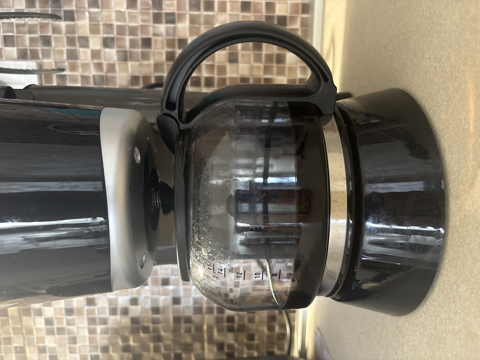 A close-up, eye-level, vertical shot shows the lower half of a black and silver coffee maker sitting on a tan countertop. The coffee maker has a glass carafe with a black handle and markings for coffee volume. The carafe contains a dark liquid, presumably coffee. Behind the coffee maker is a backsplash with a mosaic tile pattern of beige and brown squares. The scene is indoors, likely in a kitchen. No people are present.