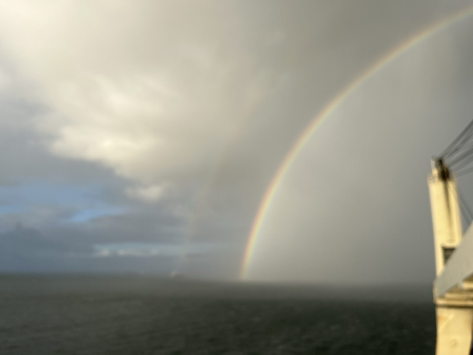 A slightly blurry image depicts a rainbow over the ocean. A second, fainter rainbow is also visible to the left of the main one. To the right, a white structural element, possibly part of a ship or building, is present. The sky is overcast, and the ocean appears dark and choppy. In the distance, a faint object that may be a ship is visible. The location is Unknown, Unknown.