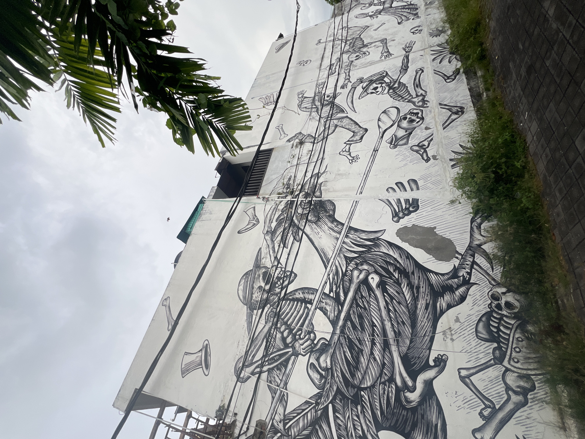 A large, intricate black and white mural dominates the side of a building in North Kuta, Indonesia, depicting numerous skeletal figures. The artwork features a prominent winged, bird-like skeletal creature in the lower section, characterized by detailed feathers made of bones and a menacing skull, seemingly engaged in a dynamic action or struggle, possibly wielding spear-like objects.
Smaller human-like skeletons are scattered across the mural, portrayed in various active poses suggesting movement, dancing, or leaping. One skeleton wears a wide-brimmed hat and holds a scythe, while another wields a large spoon-shaped object. Disembodied skulls, bones, and other abstract shapes fill the spaces between the figures, contributing to the macabre yet energetic theme.
Overhead power lines run vertically across the mural, partially obscuring parts of the artwork. Lush green palm fronds hang into the frame from the upper left, providing a natural contrast to the monochromatic mural and an overcast, light grey sky. Along the bottom edge, green grass and bushes line a paved sidewalk next to the building. The building itself is light-colored, showing some signs of wear. No visible text is present in the image.