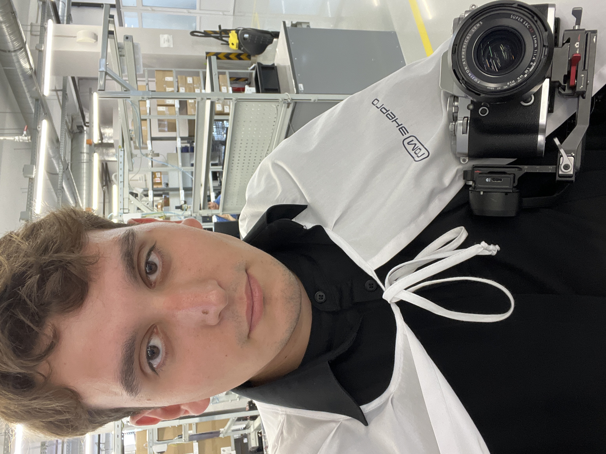 A young man is captured in a selfie-style shot, holding a camera in an industrial or laboratory environment in Zelenodol'sk, Russia. He has light brown, wavy hair and dark eyes, looking directly at the viewer with a neutral expression. He wears a black collared shirt underneath a white smock or lab coat, which has ties around the neck.
The man is holding a black and silver Fujifilm camera, specifically identified by the lens as a "FUJINON ASPHERICAL LENS SUPER EBC AF 33mm 1:1.4 R LM WR". The camera appears to be mounted within a metal cage or rig.
The background reveals a brightly lit interior space with high ceilings, featuring numerous fluorescent light tubes. Metal shelving units filled with cardboard boxes are visible, suggesting a storage or manufacturing area. There are also elements of industrial machinery, including a yellow robotic arm-like structure. The overall impression is a clean, organized factory or warehouse setting.
The white smock worn by the man prominently displays the mirrored text "ГОМ ЭНЕРГО" (GOM ENERGO) in black Cyrillic letters.