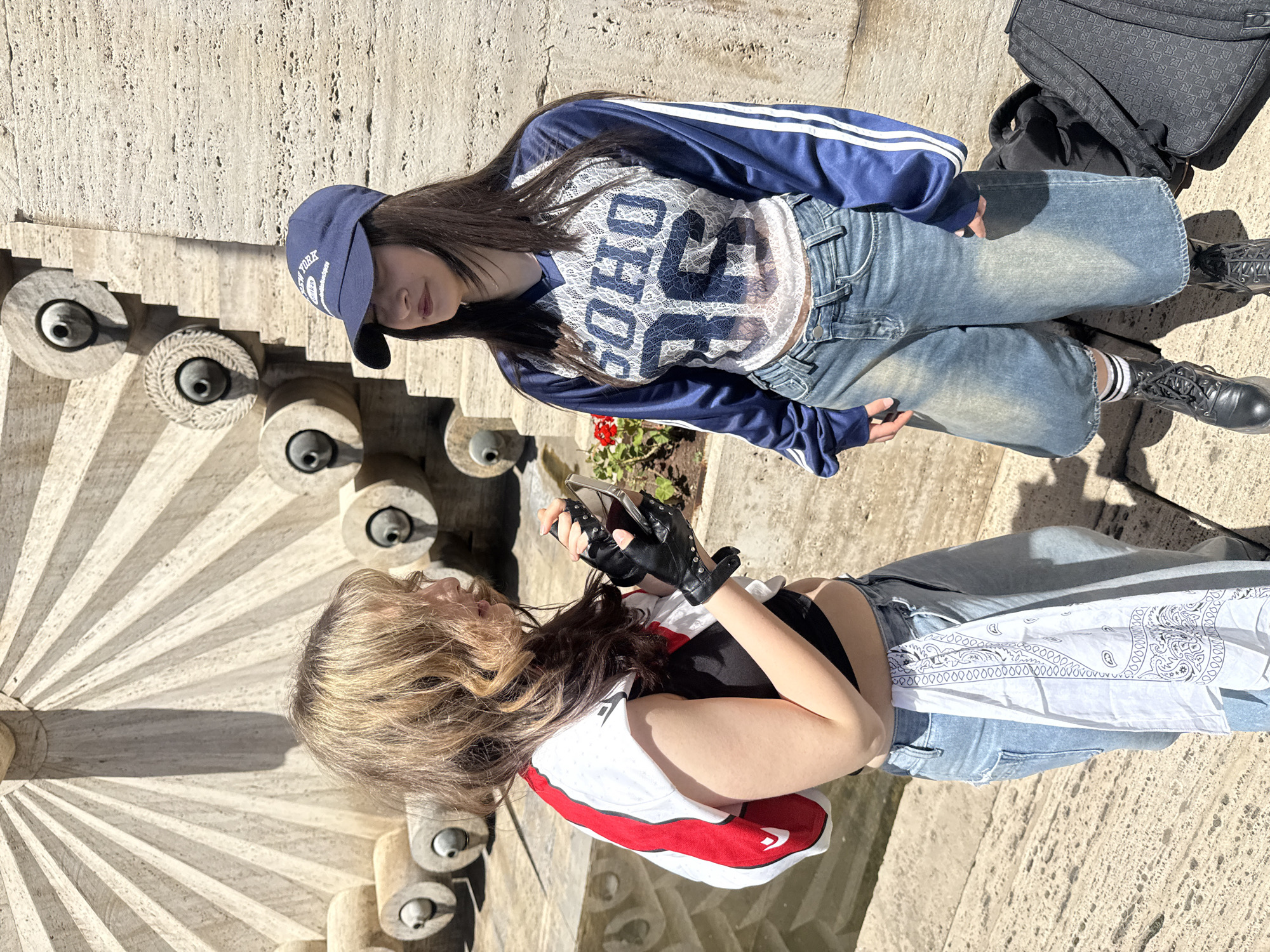 Two young women are posing for a photo on a stone structure in Yerevan, Armenia. The background suggests they are at Cascade, a monumental staircase. One woman is wearing a blue baseball cap, a blue and white top, jeans, and black boots. She is standing with a backpack next to her. The second woman is wearing a white and red jacket, a black top, jeans, and black gloves. She is holding a phone. Both are facing each other. The stone structure has a series of rounded elements. The lighting is bright.