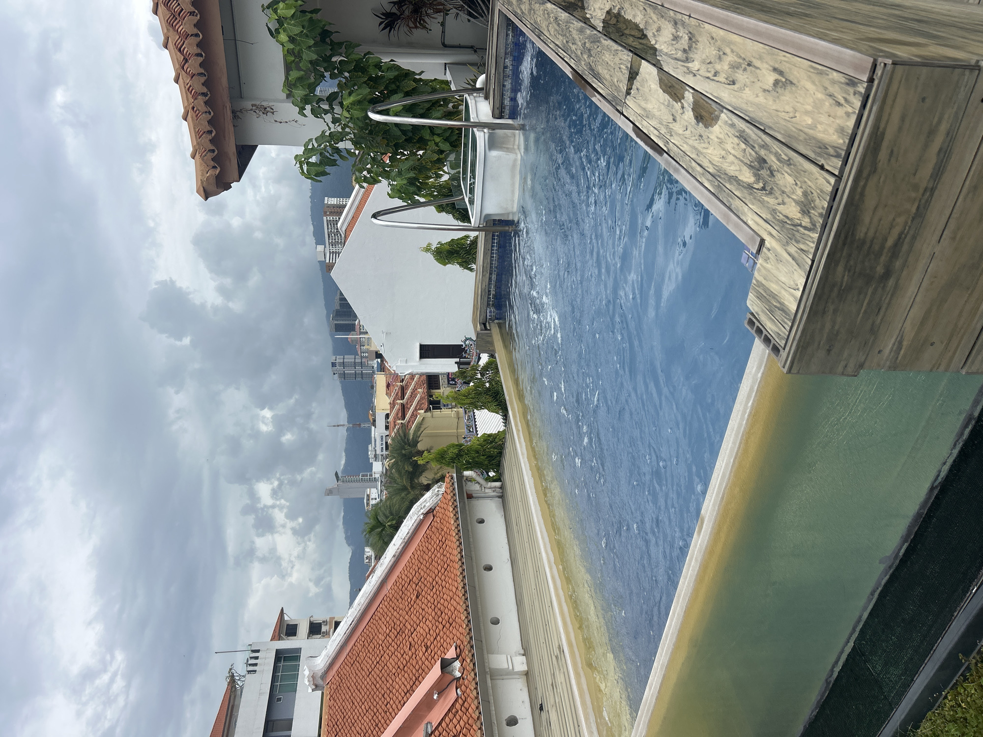 An elevated outdoor swimming pool is positioned on a rooftop in George Town, Malaysia. The pool is filled with blue water and features a built-in spa section with metal railings. The pool is surrounded by a wooden deck and a green-tinted glass barrier. In the background, a mix of traditional terracotta-tiled roofs and modern buildings are visible, with a cityscape and mountains under a cloudy sky. The image is taken from a slightly elevated angle, looking across the pool towards the cityscape.