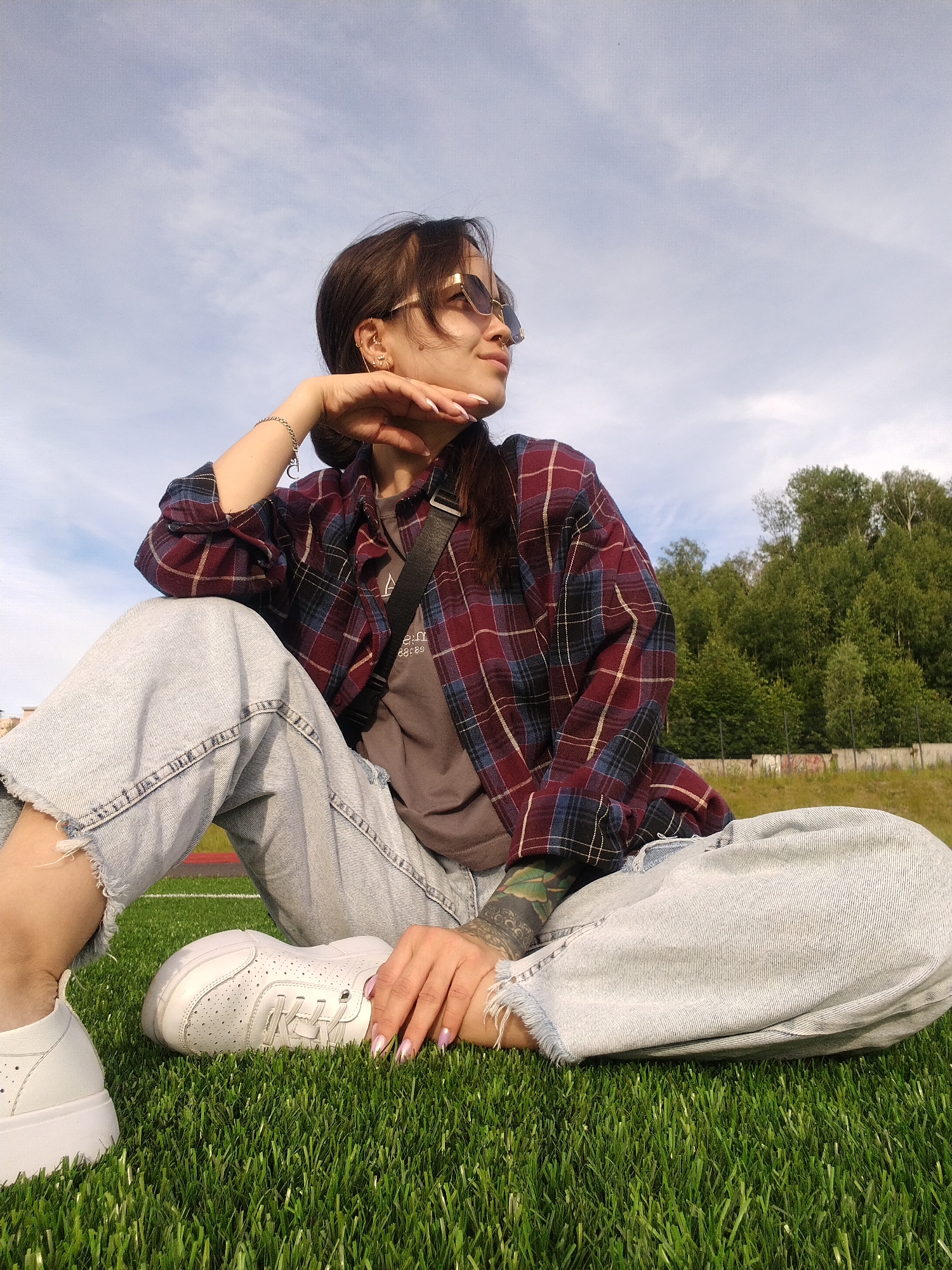 A young woman is sitting on a bright green grassy field, possibly a sports field, with a fence and a line of trees in the background. She is wearing a maroon and blue plaid shirt, a gray t-shirt underneath, and light-wash, distressed denim jeans. Her legs are crossed, and she is leaning on one hand, with her elbow resting on her knee. She is looking off to the side with sunglasses on, and her hair is pulled back. The lighting suggests it is daytime, possibly late afternoon due to the warm light and shadows. The sky is a clear blue with scattered clouds. She has tattoos visible on her left arm and a bracelet on her right wrist. The overall mood seems relaxed and contemplative.