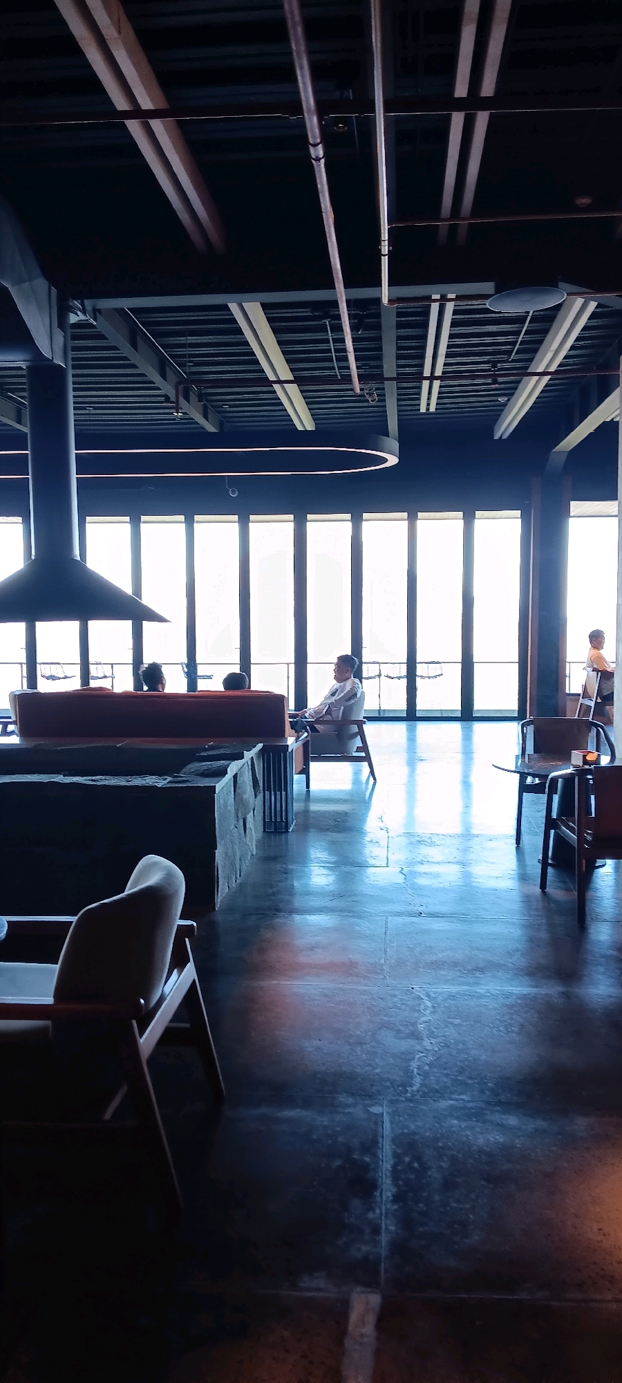 This image depicts the interior of a modern, dimly lit space, likely a restaurant or lounge, with large glass-paneled doors leading outside. The flooring is dark and reflective, showing the ambient light. In the foreground, a light-colored armchair with wooden legs is visible.
In the mid-ground, a low, dark-colored seating structure or partition is present, with a large, dark fireplace hood suspended above a section of it. Behind this, a long, orange sofa is situated, with three individuals seated on it, facing away from the camera. To the right of the sofa, a person is seated in a light-colored armchair, facing the large windows.
In the background, through the large glass doors, the exterior environment is bright and appears to be a city view, possibly Kintamani, Indonesia, given the context. The windows reveal a brightly lit outdoor scene, with some indistinct structures visible in the distance. Further to the right, another person is standing by a table, looking towards the exterior. The ceiling features exposed beams and industrial-style lighting.