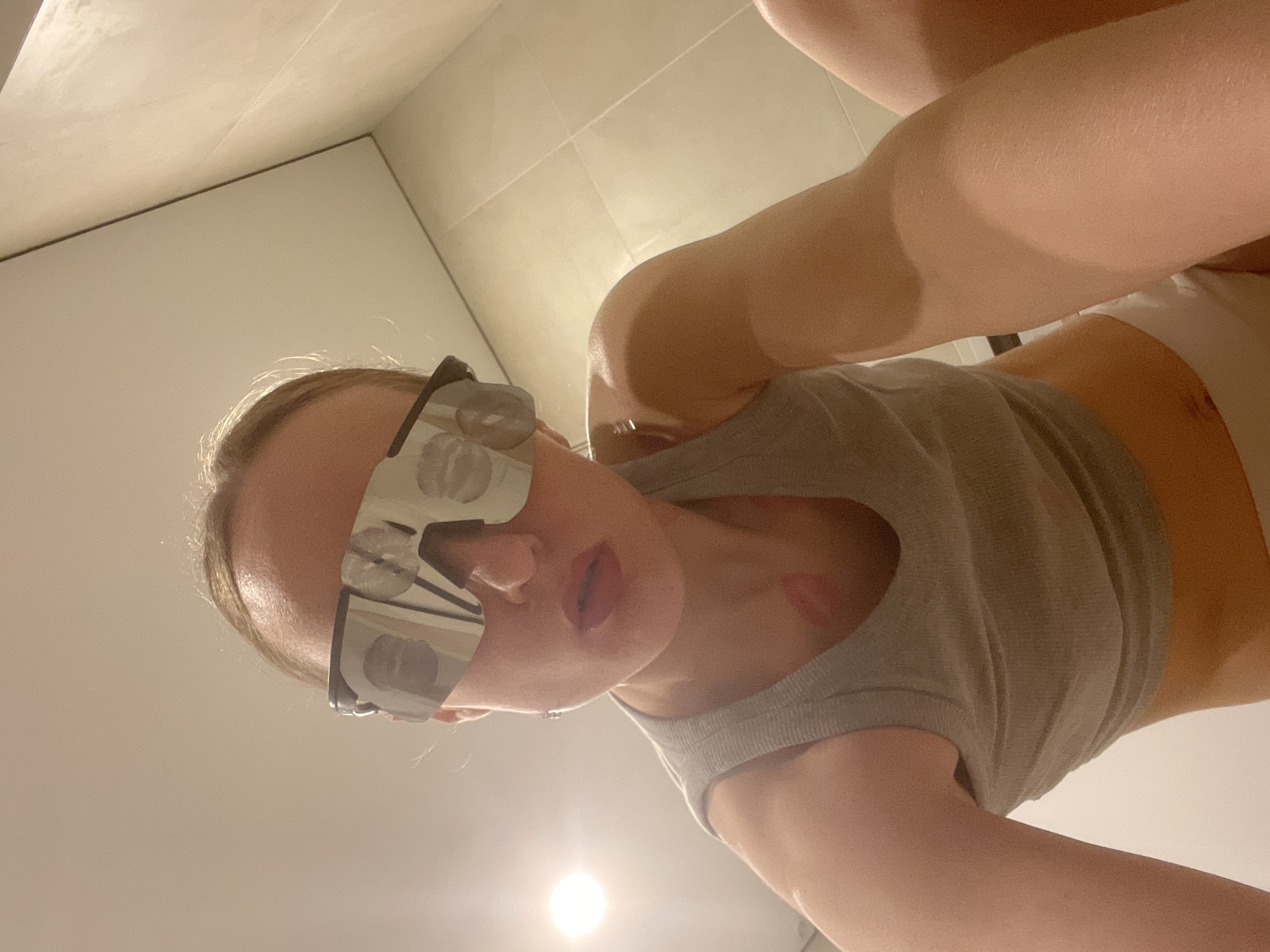 A low-angle photograph shows a woman wearing mirrored sunglasses with lipstick marks on the lenses and lipstick marks on her face and neck. She is wearing a gray tank top. The background consists of a white tiled wall and floor.