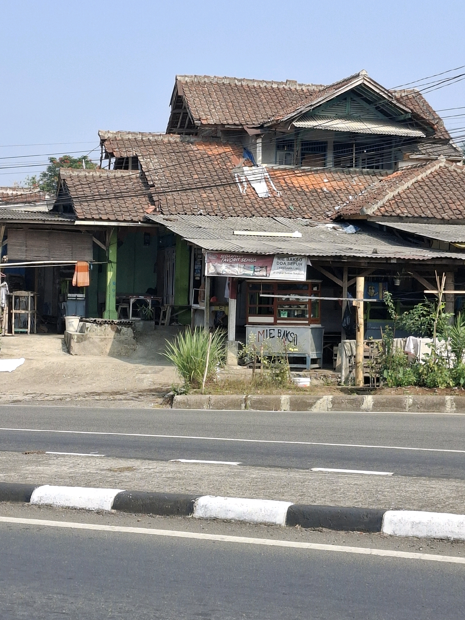 The image shows a street view in Sukabumi, Indonesia, with a row of roadside stalls and a multi-story building with a weathered, tiled roof. The building appears to be a commercial establishment or a residential building with shops on the ground floor. The stalls are simple structures with wooden frames and corrugated metal or tiled roofs, one of which displays a sign advertising "MIE BAKSO DOA SEPUH". There is a grey concrete road in the foreground with white and black painted curbing. Vegetation, including grass and small trees, is visible in front of the stalls and along the roadside. The sky is clear and blue, suggesting a sunny day. The overall scene depicts a common urban or semi-urban environment in Indonesia.