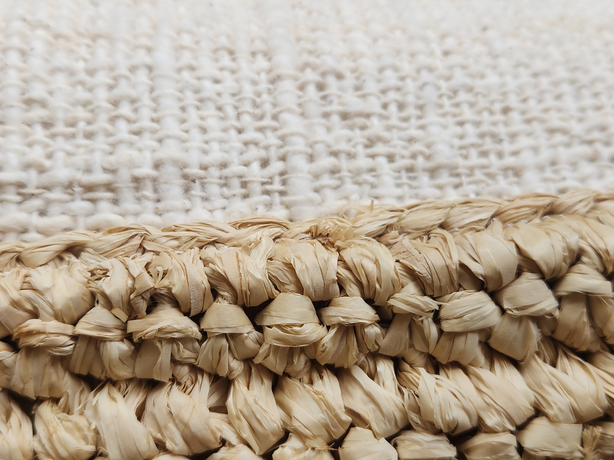 The image is a close-up, high-angle shot of a woven material. On the left side of the frame, there is a thick, textured band of woven raffia or straw in a light beige color. The strands are twisted and knotted together, creating a dense, intricate pattern. To the right of this band, the texture changes to a lighter, more loosely woven fabric. This fabric is also a light beige or off-white color and appears to have a subtle, uniform pattern of interlacing threads. The overall impression is of two different textile materials placed next to each other. The image does not contain any discernible elements that would allow for the identification of a specific location in Maitland, United States of America, or any people, actions, or interactions. The focus is entirely on the textures and patterns of the materials.