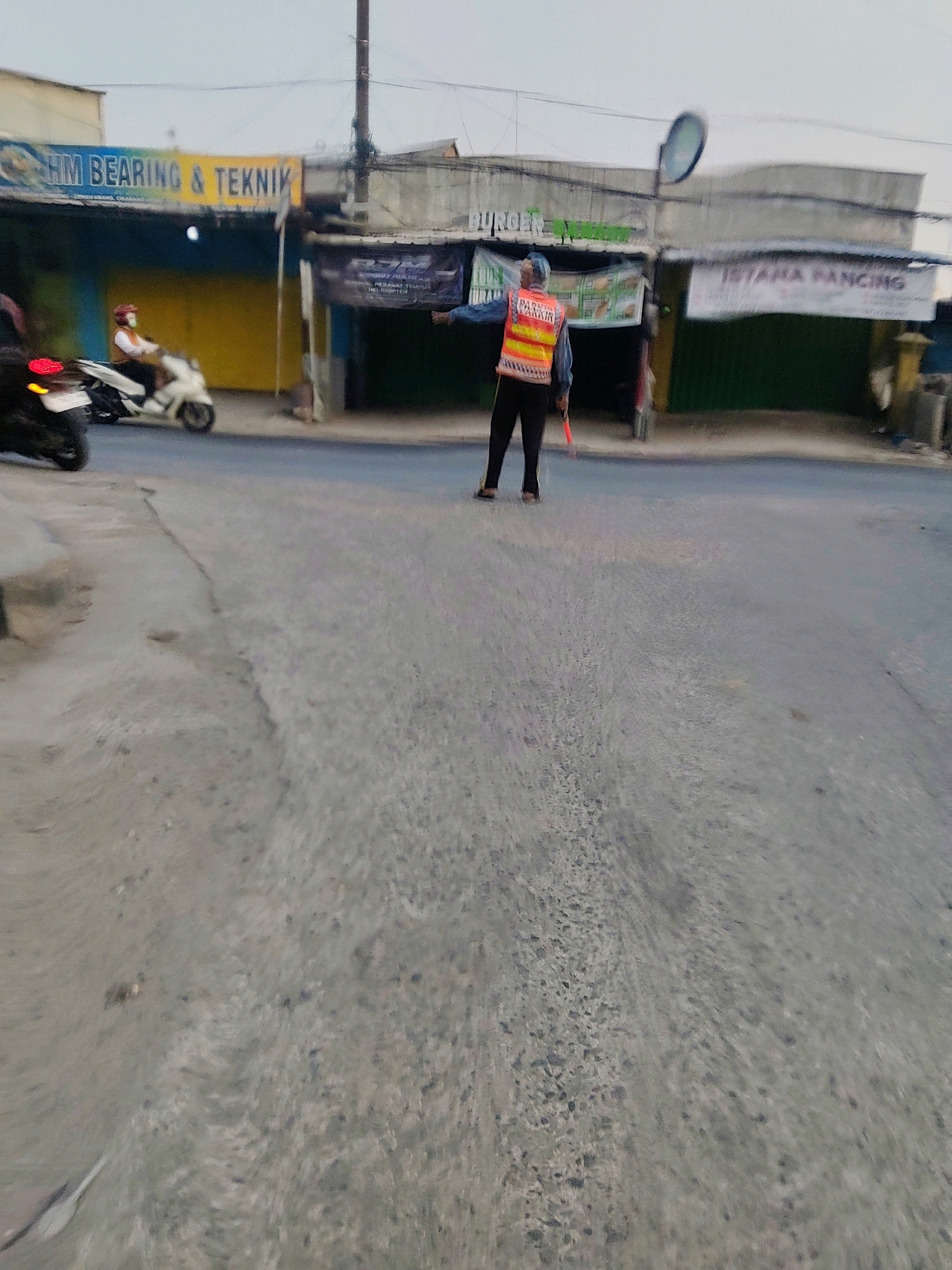 The image depicts a street scene in Mekarmukti, Indonesia, likely during the day, based on the lighting. In the foreground, a person wearing a bright orange and yellow reflective vest and dark pants is standing on the asphalt road, holding a red signaling stick. This individual appears to be directing traffic. Behind this person, a white motorcycle with a rider wearing a helmet is moving away from the camera. Along the right side of the street, several shops with signs are visible. One sign reads "ISTANA PANCING," and another banner has text that is partially obscured but appears to be related to "BURGER." On the left side of the street, a shop sign states "HM BEARING & TEKNIK." The environment appears to be a commercial or mixed-use area within a city. The road surface is asphalt, and there are some debris and cracks visible. The sky is overcast, suggesting a cloudy day.