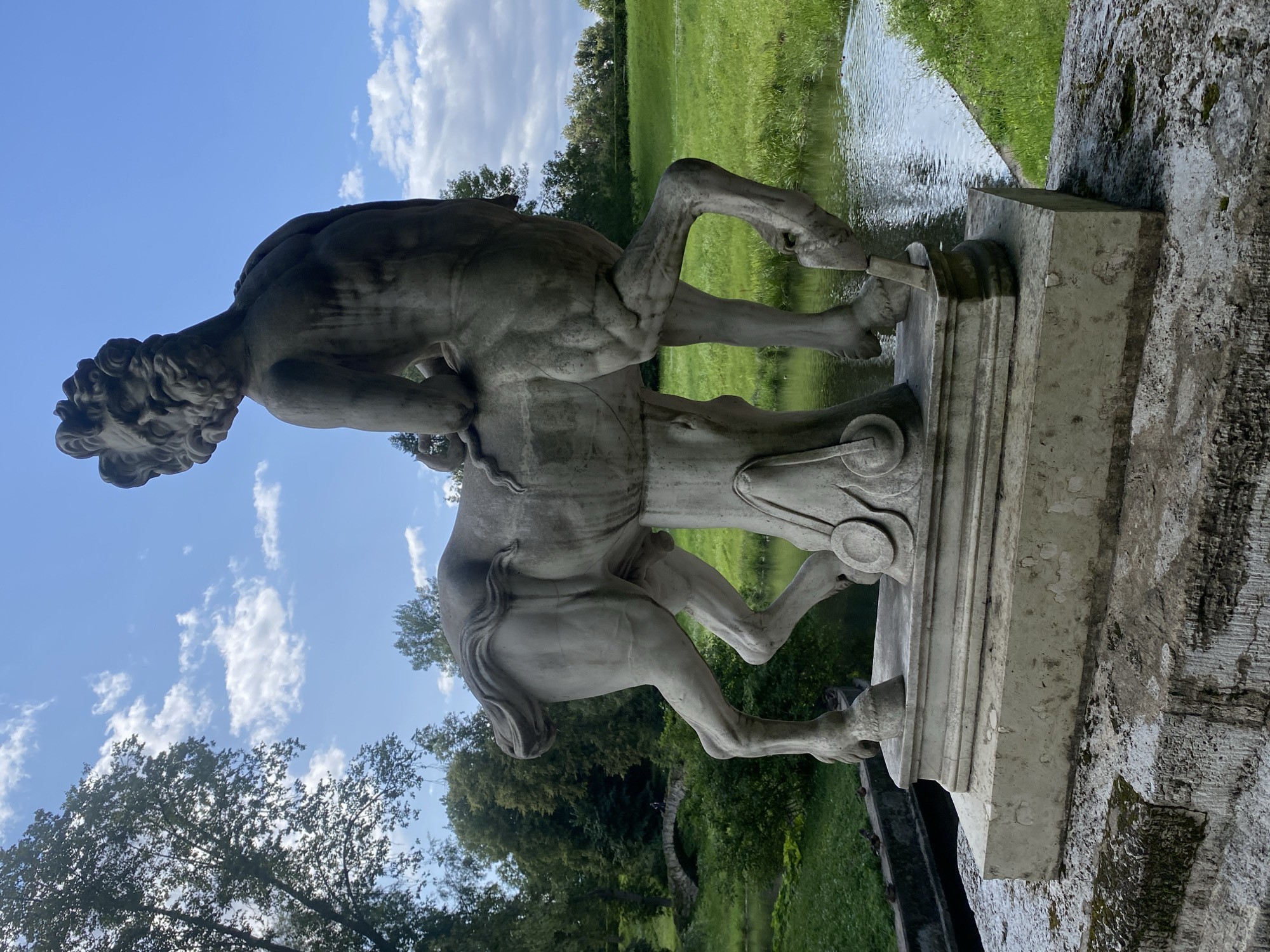 A stone sculpture of a centaur stands on a stone pedestal in the park of Pavlovsk, Russia. The centaur is depicted from the rear, with its head turned to the left. The sculpture is weathered and shows signs of age. The background consists of lush green trees, a body of water, and a stone bridge in the distance. The sky is blue with scattered white clouds. There are no people visible in the immediate vicinity of the sculpture. The overall scene evokes a sense of classical beauty and tranquility.