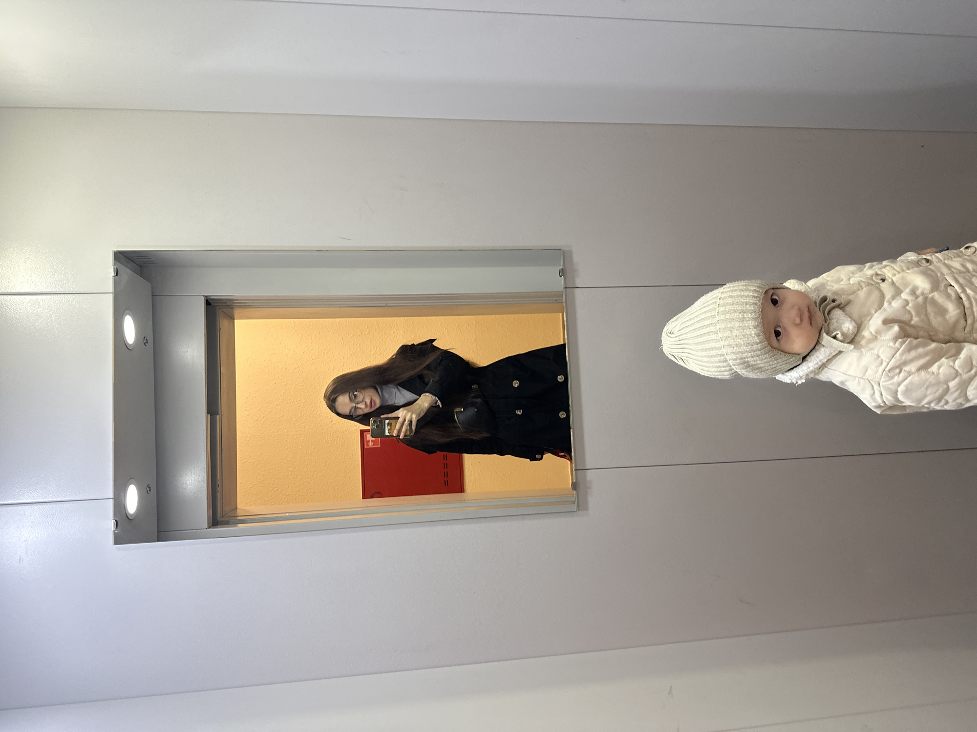 The image depicts a woman taking a mirror selfie in what appears to be an elevator. The woman is wearing a black coat and glasses, holding a phone. A baby is present, dressed in a white quilted snowsuit and a knit cap. The mirror reflects an interior space, possibly an office or building lobby, featuring a red fire hose cabinet on an orange-painted wall. The elevator is located in Yekaterinburg, Russia.