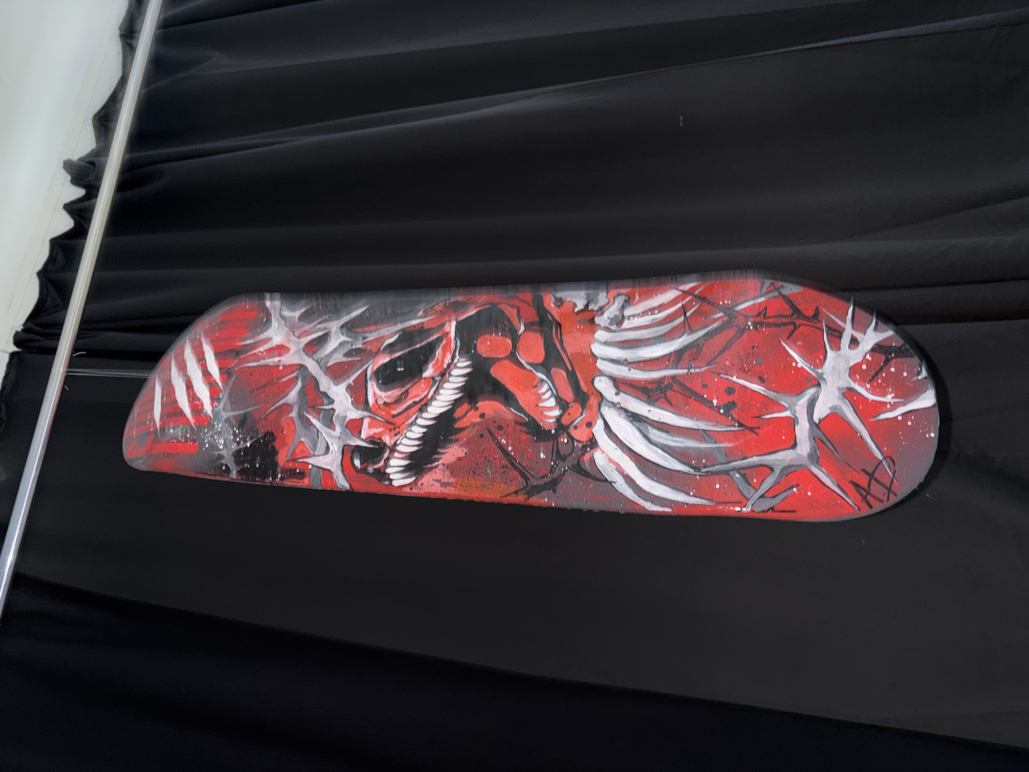 The main subject of the image is a skateboard deck featuring a vibrant and intense artwork, displayed against a dark, textured background.
The skateboard deck is adorned with a custom design predominantly in red, black, white, and grey. The artwork depicts a stylized, skeletal, or monstrous face on the left side, integrated with jagged, rib-cage-like structures and sharp lines that extend across the length of the board. The color scheme is aggressive, with a bright red base providing a stark contrast to the white and grey skeletal elements and the black outlines and shading. Splatter effects and fine lines add texture and depth to the graphic, creating a dynamic and somewhat gothic or street art aesthetic.
The skateboard is positioned horizontally, slightly angled, against a backdrop of dark, possibly black, draped fabric that exhibits gentle folds and creases. A shiny, vertical metal pole or rail is visible along the left edge of the frame, suggesting an indoor setting, possibly an art exhibition, shop display, or a studio. The overall scene implies the skateboard is being presented as an artistic object rather than being in active use.
There is no activity taking place; the image captures a static display. Notable details include the aggressive and intricate design on the skateboard, with its strong visual impact. Faint, intertwined markings, possibly a signature or initials like "AX" or "AR", can be discerned within the white skeletal pattern on the far right end of the board. The lighting appears even, suggesting an indoor environment without strong directional light that would indicate a specific time of day. The location context provided is Krasnoyarsk, Russia.