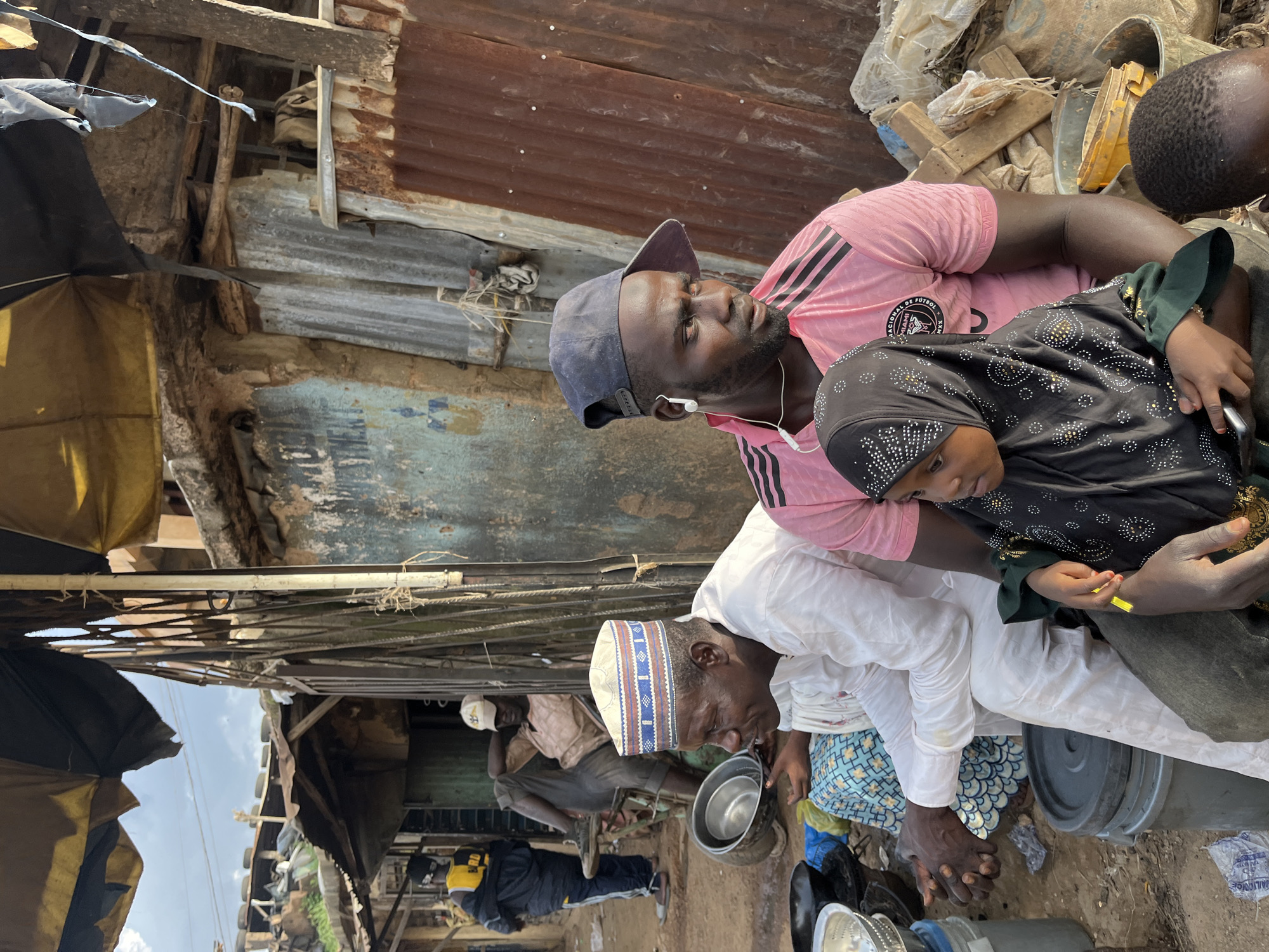 In the city of Rafin Par, Nigeria, three individuals are captured in a medium shot, seated outdoors amidst a chaotic environment. The foreground is dominated by a man wearing a pink t-shirt and a dark baseball cap, with white earbuds in place. To his right, a young girl in a black hijab is seated on his lap, her hand resting on his arm, holding a yellow object. To the left of the man in the pink shirt, another man in a white traditional outfit and a patterned cap is seated. His hands are clasped in his lap, and he is looking to his left. Behind them, other people are visible in the background, including a man sitting on a bicycle and another standing further back wearing a yellow and black jacket. The environment appears to be a market or a bustling street, with makeshift structures constructed from corrugated metal and fabric. Piles of discarded materials, buckets, and metal bowls are scattered around the scene, suggesting a working-class or informal settlement. The overall impression is one of everyday life unfolding in a densely populated urban setting.