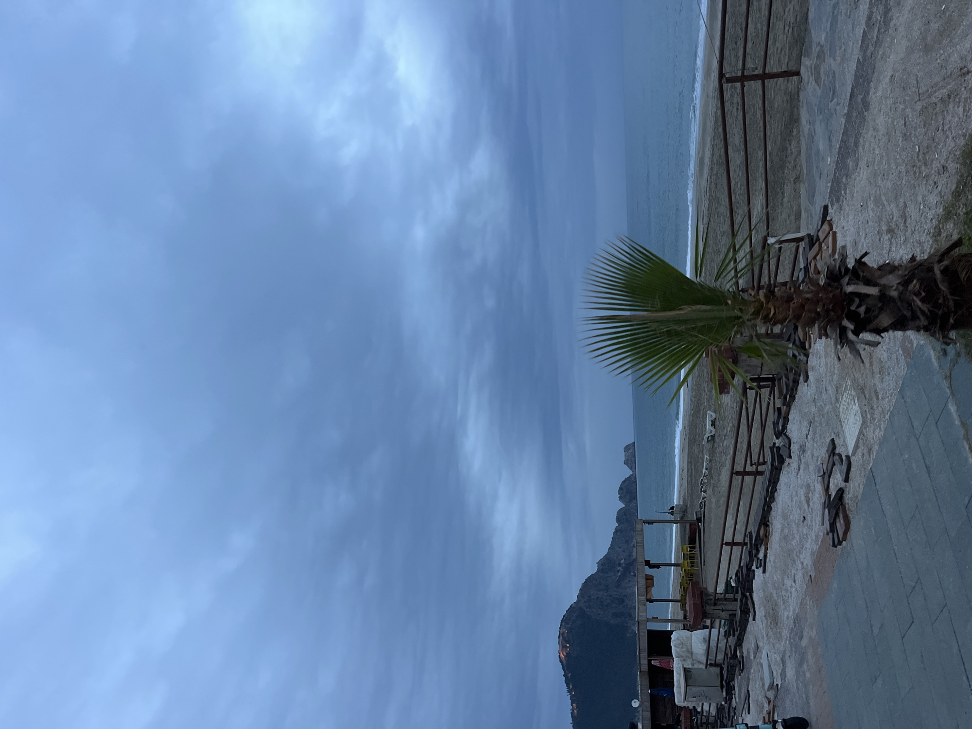 The image shows a view from a beach in Alanya, Turkey. A palm tree is visible in the foreground along the railing and sidewalk. The scene includes the beach, the ocean, a mountain, and a cloudy sky. Construction materials and restaurant tables and chairs are visible. The photograph has been rotated.