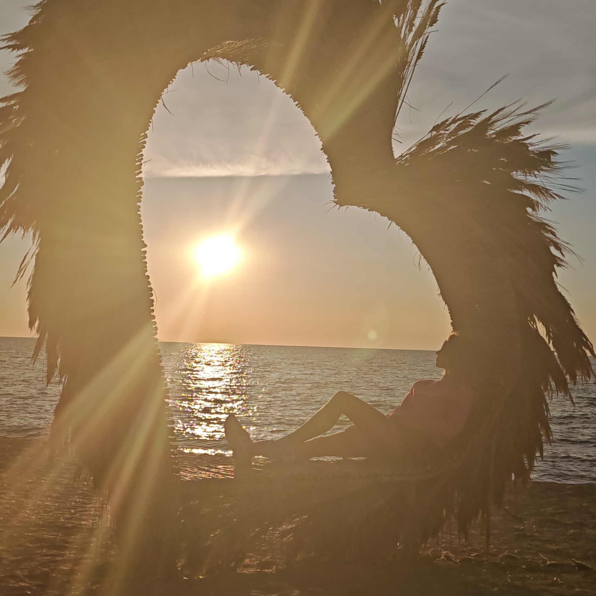 A person is reclining on a beach, silhouetted against a sunset. The person is positioned within a large, heart-shaped structure made of natural materials, possibly palm fronds, which frames the view of the ocean and sky. The sun is low on the horizon, casting a warm, golden light and creating a shimmering reflection on the water. The sky is a soft gradient of yellow and orange, with some wispy clouds. The beach appears to be made of dark sand. The overall scene evokes a sense of peace and relaxation.