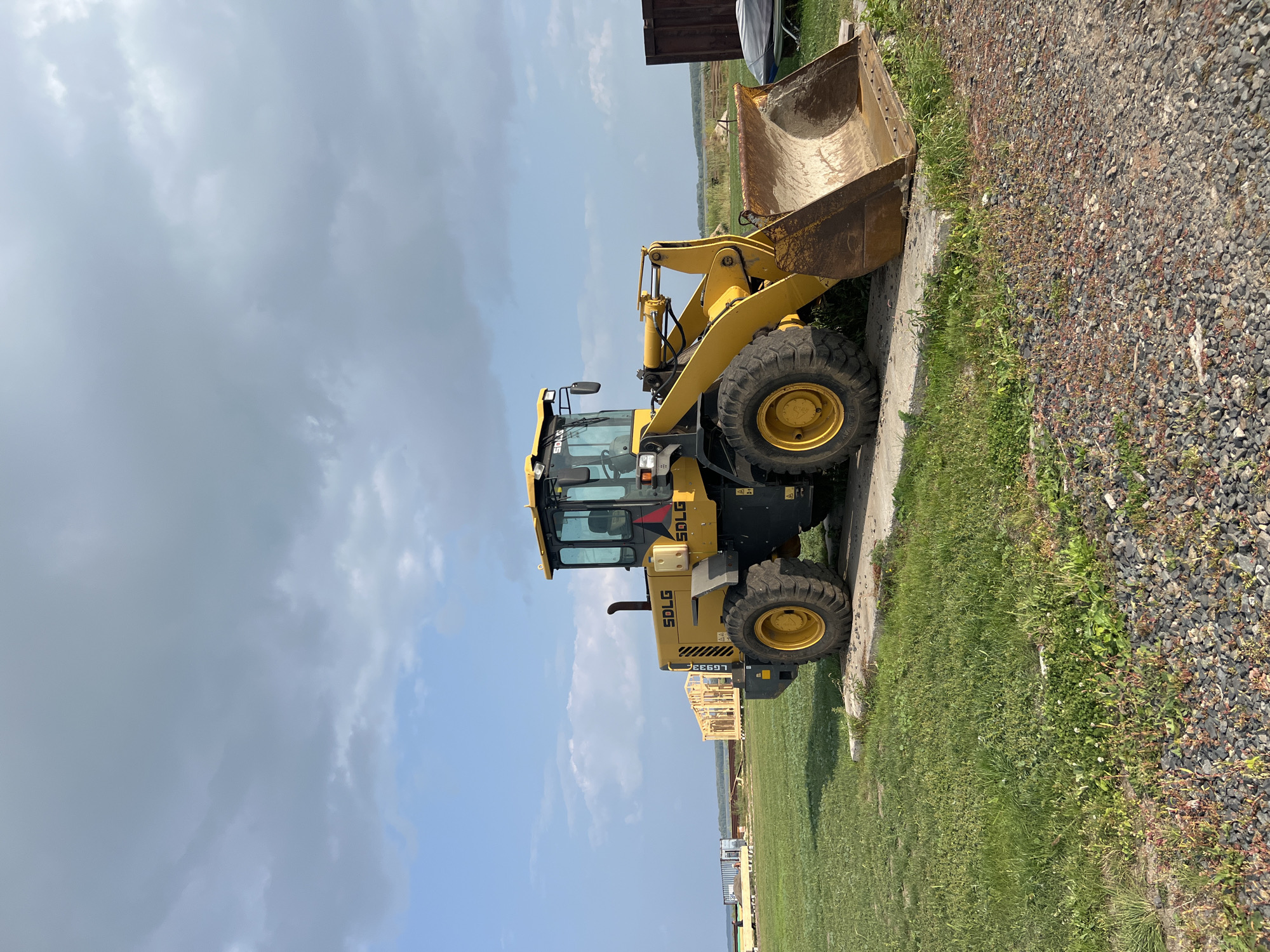 An eye-level, medium shot captures a yellow SDLG wheel loader parked on a concrete pad in an outdoor, grassy area. The loader, facing slightly to the left, is positioned in the foreground, its bucket extended. The background reveals a construction site with wooden framing for structures, a body of water in the distance, and a cloudy sky overhead. The scene is likely located in Novopichugovo, Russia, given the context provided. The environment is rural or semi-rural, characterized by open fields and construction activity under a dynamic sky. No people are visible in the image.