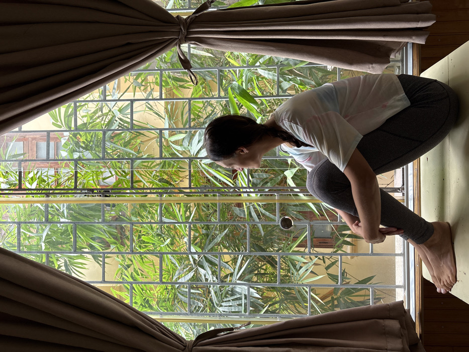 An adult female, positioned in profile facing right, is seated barefoot on a light-colored surface, possibly a window sill or low bench, within an interior space. She wears a white short-sleeved t-shirt with subtle pastel markings and dark gray patterned leggings. Her dark hair is styled in a braid. Her gaze is directed outwards through a large window, framed on either side by tied-back brown curtains. The window is secured by a prominent gray metal grid of rectangular bars. Beyond the window and security bars, dense green foliage, consistent with bamboo, is visible, partially obscuring a light-yellow building with dark-framed windows in the background. The environment, characterized by dense urban greenery and visible architecture, is consistent with a residential or guesthouse courtyard common in the city of Hà Nội, Vietnam.
