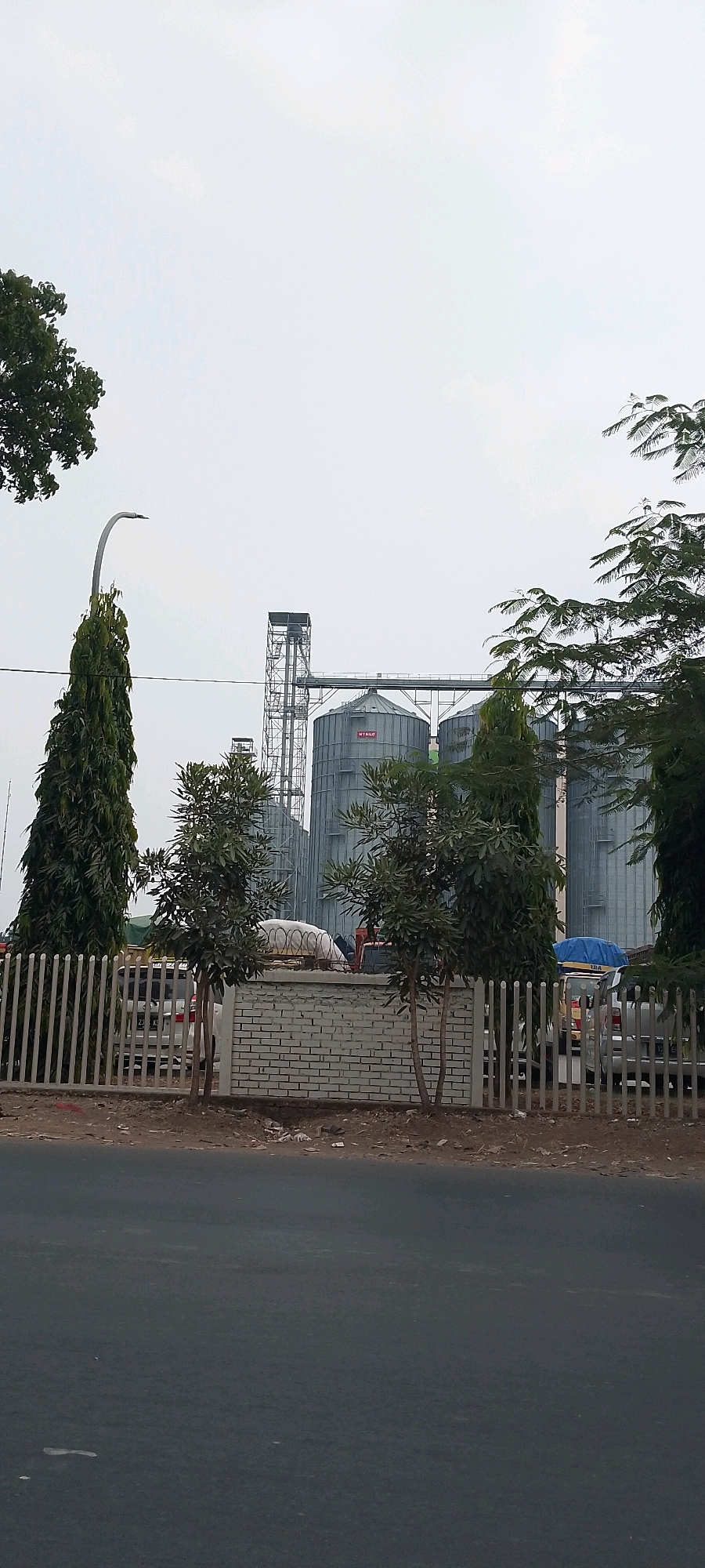 The image shows industrial silos in Bekasi, Indonesia, under a pale sky. In the foreground, a gray asphalt road is visible, with a dirt shoulder littered with trash. A white metal fence runs along the edge of the property. Behind the fence, a low white brick wall separates the road from the industrial area.
Several large, cylindrical gray silos are the dominant features of the industrial complex. A metal structure with a walkway connects the tops of some of the silos, and a tall, narrow metal tower stands to the left of the silos.
Trees are scattered throughout the scene. Two tall, slender evergreen trees stand to the left of the silos, and a street lamp is positioned between them. To the right of the silos, a dense cluster of leafy trees partially obscures the view of the industrial structures.
Several vehicles are visible behind the fence. A white car is parked on the left side, and a vehicle with a blue tarp is to the right. A person is partially visible near a pile of what appears to be bagged material behind the brick wall. The ground behind the fence is a mix of dirt and some vegetation.