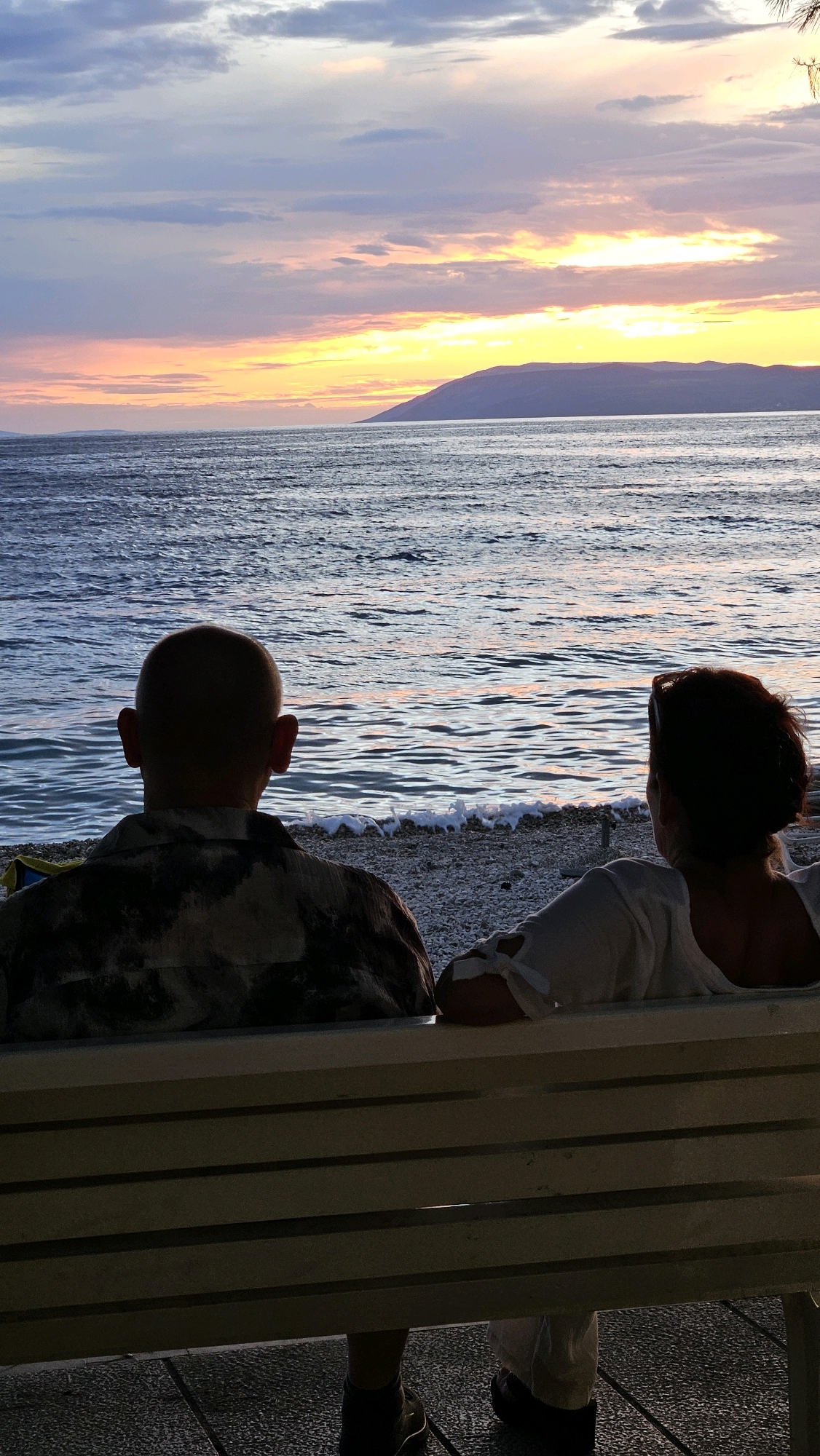 Two individuals, seen from behind, are seated on a bench facing a body of water and a distant coastline during sunset. The person on the left has a shaved head and is wearing a patterned shirt. The person on the right has dark hair pulled back and is wearing a white shirt. The bench is light-colored with horizontal slats, situated on a paved area next to a pebble beach. The water is calm with gentle waves, reflecting the colors of the sky. The sky displays a gradient of orange, yellow, pink, and purple hues, indicative of a sunset, with clouds scattered across it. A silhouetted landmass, likely an island or mountain range, is visible on the horizon. This scene is located on the coast of Općina Tučepi, Croatia.