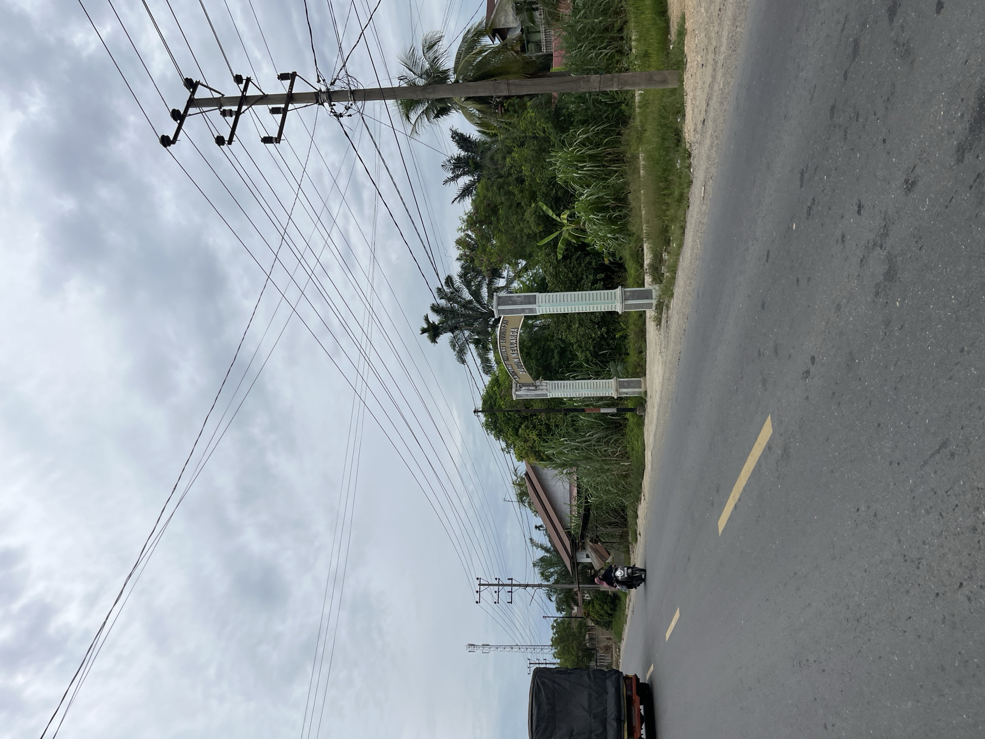 This image captures a scene on a cloudy day in Simpang Empat, Indonesia. A paved road runs through the frame, with a yellow dashed line marking the lane. On the left side of the road, a large truck with a black tarp covers its cargo is moving away from the camera. Further down the road, a person is riding a motorcycle.
On the right side of the road, a concrete archway stands with text that appears to read "KAMPUNG TANDUNG SEPULUH" and "DUSUN KELUARGA". This archway is flanked by dense vegetation, including tall grasses and palm trees, and behind it, houses with brown roofs are visible. A utility pole with numerous wires crisscrossing overhead stands prominently on the right side of the road, near the archway. In the distance, another utility pole and a cell tower are visible. The sky is overcast with gray clouds.