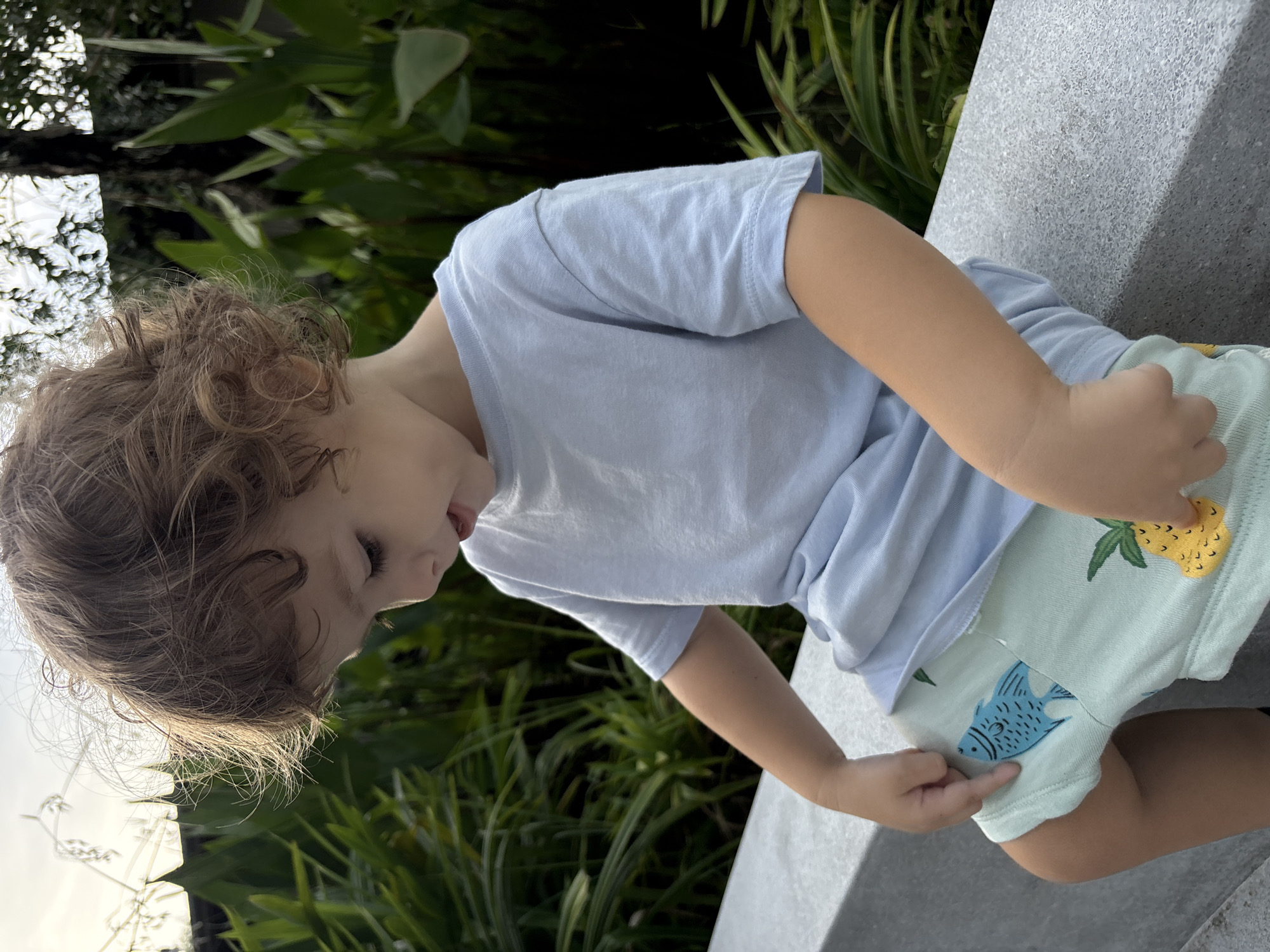 A young child is seated outdoors on a light gray concrete or stone bench in Choeng Thale, Thailand. The child, presenting with light brown, curly hair and fair skin, is wearing a plain light blue short-sleeved t-shirt and light green shorts. The shorts feature a distinct blue fish graphic on the upper left thigh and a yellow pineapple graphic on the upper right thigh. The child is looking downwards and slightly to the left, with the mouth slightly open. The hands are positioned near the thighs; the right index finger points towards the blue fish graphic on the left thigh. The background consists of dense green tropical foliage, including broad-leafed plants and thinner, spiky vegetation. The upper portion of the background reveals a bright, hazy sky with hints of structural elements and a dark tree trunk on the right. The scene is illuminated by natural daylight, casting some light onto the child's hair from the upper left.