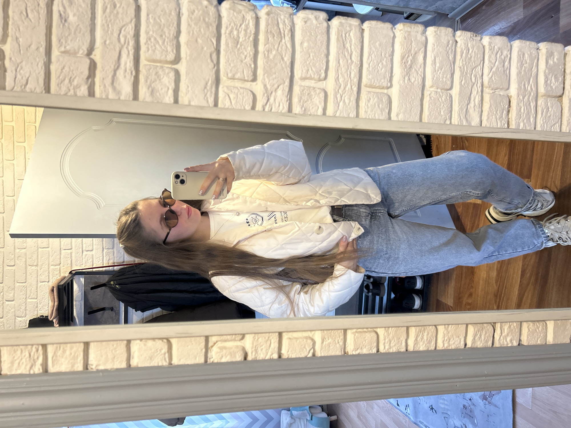 A female individual is depicted standing full-length in a mirror, capturing a selfie with a light-colored smartphone held in her right hand. She wears brown-tinted oval sunglasses, a white quilted jacket over a white long-sleeved t-shirt displaying reversed text including "KLIN" and "MOSCOW", light-wash baggy denim jeans, and chunky white and grey sneakers. A dark shoulder bag strap is visible. The reflection reveals an indoor residential setting with white brick-effect walls, a white paneled door with an arched top design, and light brown wooden flooring. To the left, a blue and white patterned rug is on the floor, and a piece of light blue and white striped furniture is partially visible. Behind the subject, a black metal multi-tiered shelf unit holds items including a dark bag, folded textiles, and a dark jacket on a hook. The scene is located in the city of Klin, Russia, as identified by the text on the subject's apparel.