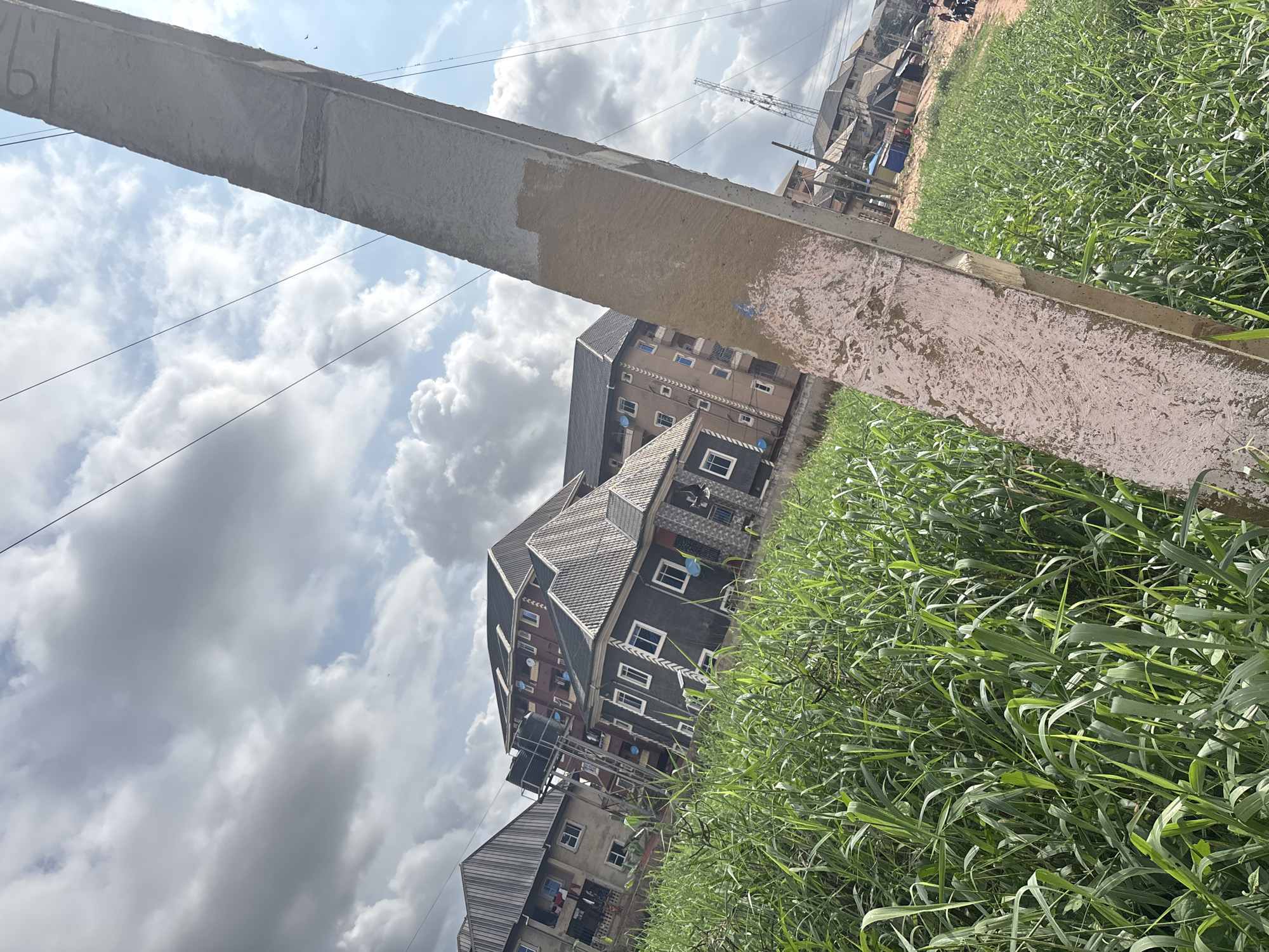 This is a slightly tilted, eye-level shot of a grassy field with buildings in the background under a partly cloudy sky, likely in Nkpor, Nigeria. In the foreground, a concrete utility pole, weathered and with patches of pink paint, is positioned diagonally across the right side of the frame. The pole is rough in texture and shows signs of age.
To the left of the pole, a dense field of tall, green grass dominates the lower half of the image, reaching up towards the buildings. The grass appears healthy and lush.
In the background, several multi-story residential buildings are visible. The closest building on the left is a lighter-colored structure with visible windows and balconies. To its right, a darker, modern-looking building with a grey roof and white-framed windows is prominent. Further back, other buildings of varying colors and architectural styles are present, including one with a water tank on its roof and a reddish-brown facade. In the far background, a communication tower can be seen.
The sky is a mix of blue and grey clouds, with sunlight breaking through in some areas. The overall impression is of a residential area on the outskirts of a city, with a mix of developing infrastructure and natural elements.