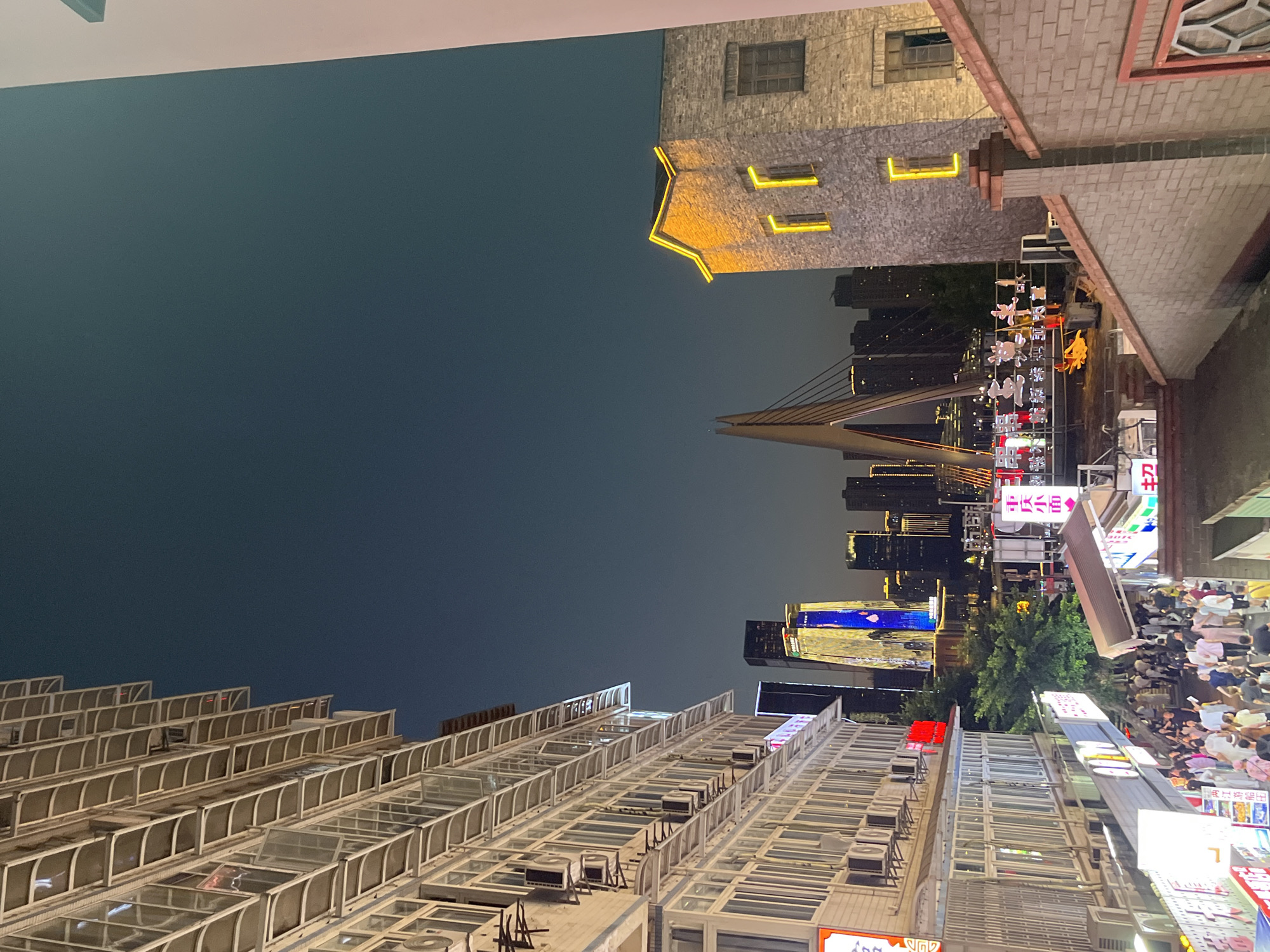 This is a nighttime photograph taken in the Yuzhong District of Chongqing, China, likely from an elevated vantage point. The image captures a bustling street scene below a modern cityscape.
In the foreground, a multi-story building with numerous windows and air conditioning units is prominently featured on the left side of the frame. The building has a light-colored facade with white window frames and awnings.
To the right, a brick building with a distinctive peaked roof illuminated by yellow lights is visible. This building has small, dark windows. Beyond it, a section of a red-tiled roof with a decorative element is also in view.
The middle ground reveals a densely populated pedestrian area with people walking and congregating. Various signs with Chinese characters are displayed above the shops. A distinctive bridge with a tall, modern pylon and cables spans across the water in the distance, illuminated by lights.
The background showcases a sprawling cityscape with numerous tall buildings, some of which are lit up, including a prominent skyscraper with vertical blue and yellow lighting. The sky is a deep, dark blue, indicating nighttime. The overall atmosphere suggests a lively urban environment.
