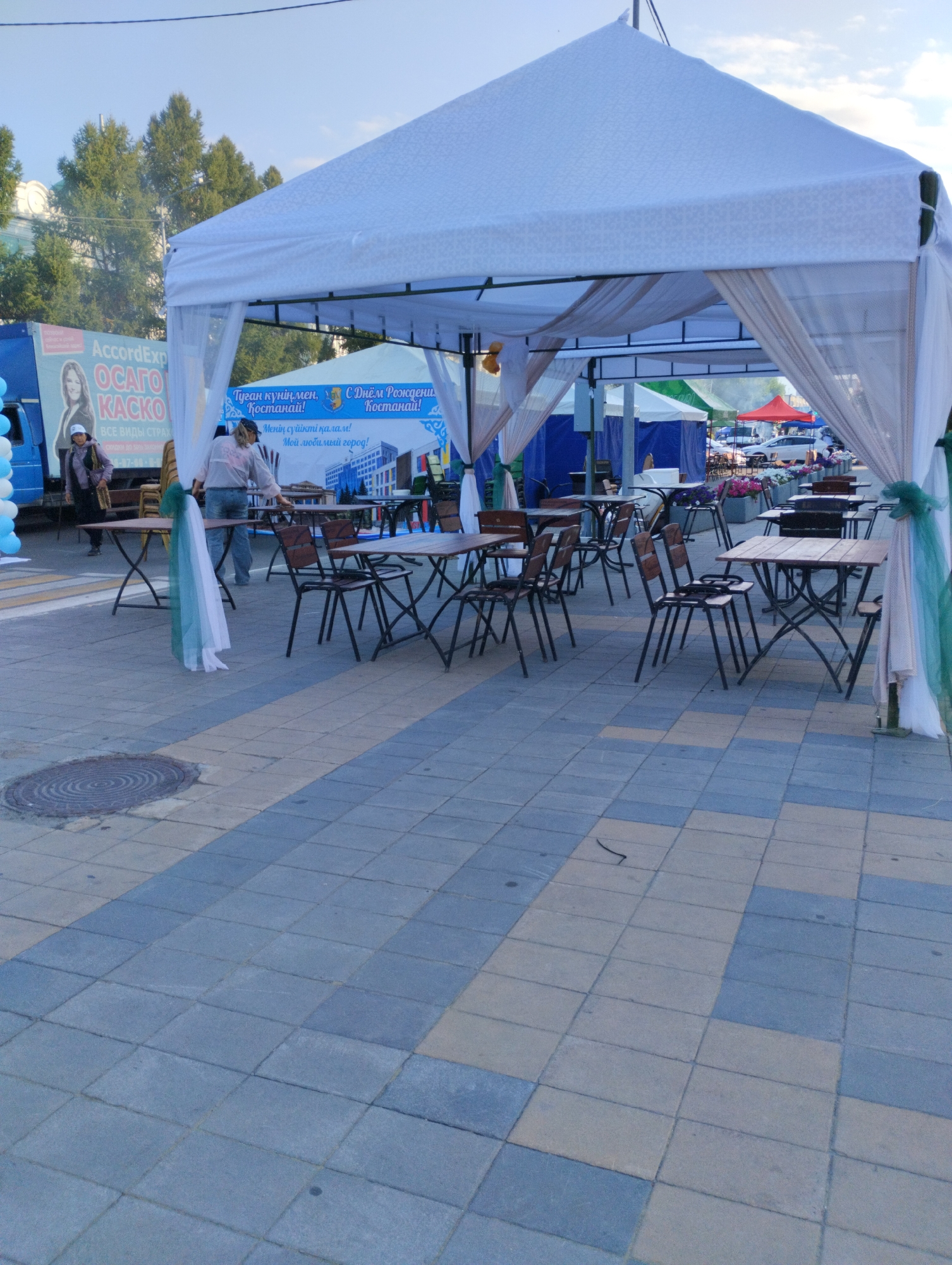 The image depicts an outdoor market or event set up under a large white tent with draping fabric. Several tables and chairs are arranged on a paved walkway, some of which appear to be made of wood with metal frames. In the background, there are other tents, vehicles, and trees. A blue truck with signage for "AccordExp OCAТO KACKO" is visible on the left side of the image. A banner with text in Kazakh and Russian, including "С Днём Рождения, Костанай!" (Happy Birthday, Kostanay!), indicates the location. Two individuals are present in the scene: one standing near the truck and another near the tables. The ground is paved with rectangular tiles in alternating shades of grey and blue. A circular manhole cover is visible on the pavement in the lower left quadrant. The overall scene suggests a public gathering or market in the city of Kostanay, Kazakhstan.
