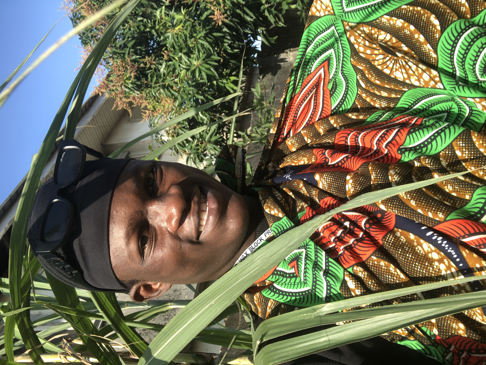The photograph is a close-up, outdoor, portrait-style shot of a young Black man standing amongst large green leaves, possibly sugarcane, in Ado, Nigeria. He is wearing a black beanie with black sunglasses perched on top. His attire features a vibrant, colorful dashiki with a geometric and floral pattern in shades of green, red, orange, blue, and brown. He is smiling at the camera, revealing white teeth. Behind him, out of focus, is a building with a white roof and a wall, and lush green foliage, likely trees, with some reddish-brown blossoms or fruits. The sky above is a clear, bright blue. The overall impression is a sunny, warm day.