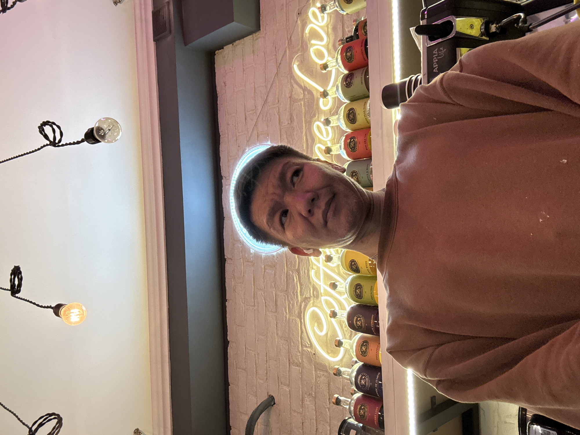 This media file is a selfie taken by a man in an indoor setting, possibly a bar or restaurant, as indicated by the presence of numerous bottles displayed on shelves in the background. The man is wearing a brown sweatshirt and is positioned in the foreground, looking directly at the camera. Behind him, a neon sign spells out "the Love" in yellow against a white brick wall. The bottles on the shelves appear to be various liquors or liqueurs, with labels visible, some of which are partially legible, showing text such as "KONG," "CURRANT," "PISTACHIO," "PINEAPPLE," and "RED ORANGE." There are also some decorative light bulbs hanging from the ceiling in the upper left portion of the image, casting a warm glow. The overall lighting suggests it could be evening or a dimly lit establishment. The man's expression is neutral. The text "APPIA Life" is visible on a black object in the upper right. The context provided indicates the location is Moscow, Russia.