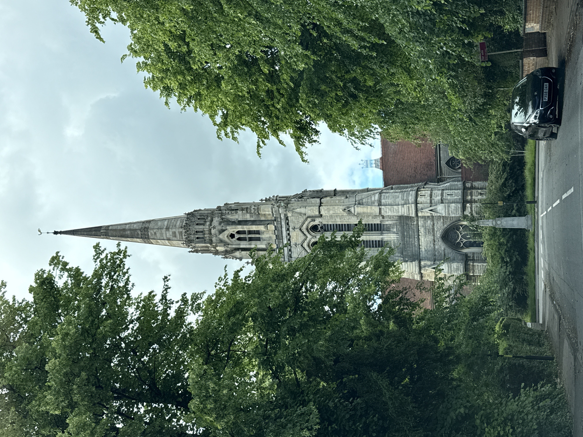 The image displays a tall, ornate stone church spire and tower, captured from a street-level perspective. The church, exhibiting intricate Gothic architecture, dominates the central frame, rising dramatically against an overcast, cloudy sky. Detailed carvings, lancet windows, and various architectural elements are visible on the gray stone structure, with the spire culminating in a sharp point topped by a finial.
Lush green trees frame the scene, with foliage partially obscuring the church on both sides and in the foreground. To the right of the church, a paved road with a dashed white line is visible. A dark-colored car, likely a hatchback, is parked on the side of this road, facing away from the viewer. The car's rear license plate, "PX68 OAH," is clearly legible. Above the car and partially hidden by trees, a red "For Sale" sign is visible on a pole. Part of another brick building, possibly an annex to the church or an adjacent structure, can be seen through the trees on the right.
The scene depicts a quiet daytime setting under an overcast sky, suggesting soft, diffuse light. No people are present, and there is no apparent activity other than the stationary car. The combination of the grand church, dense greenery, and road with a parked car suggests a setting in a town or city, likely in the UK given the license plate format.