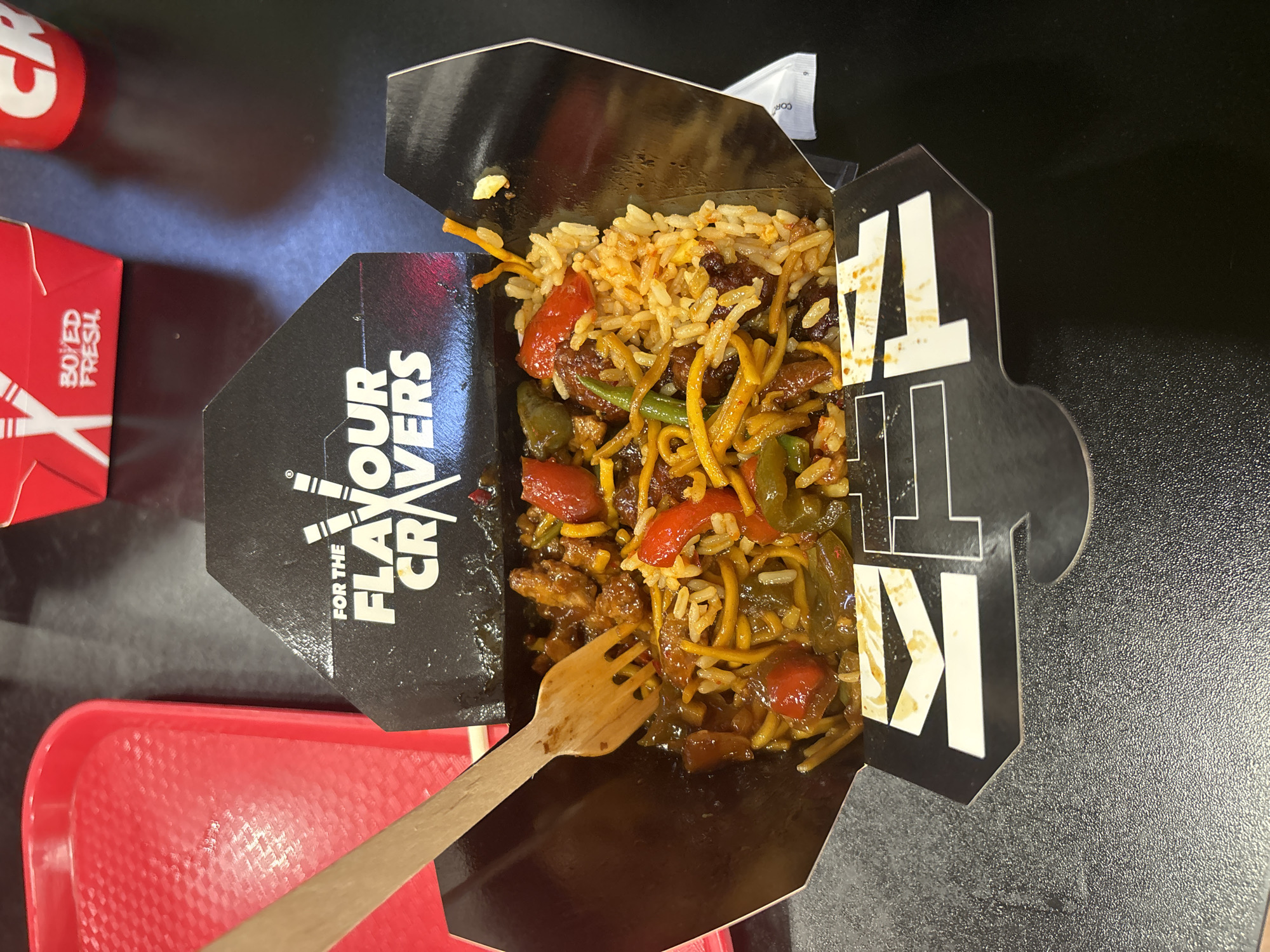The image shows a close-up, top-down view of a takeaway box filled with food, likely noodles and rice with vegetables and meat. A wooden fork is positioned in the bottom left of the box, about to pick up some food. The box is black with white text that reads "FOR THE FLAVOUR CRAVERS" and also features large white letters spelling out "TA K I". The food appears to be a stir-fry dish with visible pieces of red bell pepper, green beans, and meat. Some of the food has spilled slightly onto the black interior of the box. To the left of the food box, a red tray is visible, and further to the left, parts of another red takeaway container with white text that reads "BO ED FRESH" are visible. The setting appears to be a table, likely in a casual dining environment or at home. The lighting suggests it could be daytime. No people are visible in the image.