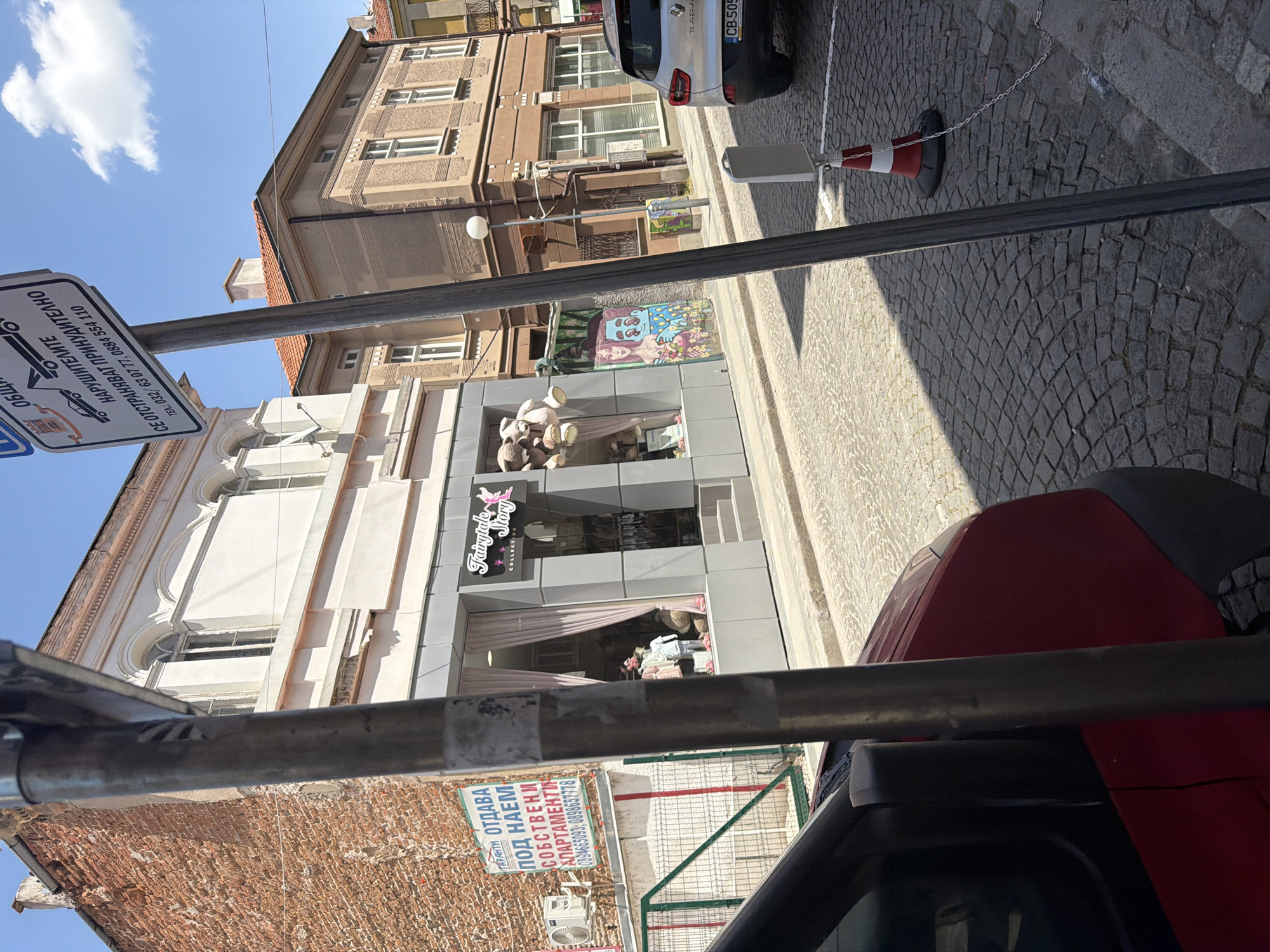 The image captures a street scene in Plovdiv, Bulgaria. The primary subjects include buildings, a street sign, vehicles, and street furniture. The "Fairytale Story Collection" store is visible, indicating retail activity. An adjacent building has a sign advertising apartments for rent, suggesting a mix of commercial and residential properties. The street is paved with cobblestones, and a parking sign indicates parking regulations. A red car is partially visible in the foreground. The weather appears sunny, with a clear blue sky.