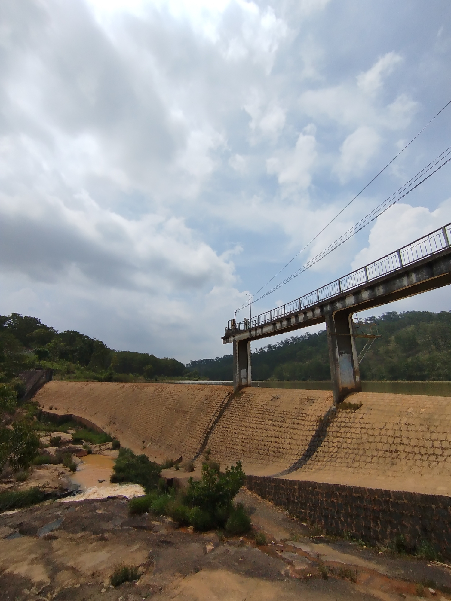 This wide-angle shot depicts a dam or weir structure set against a backdrop of rolling, tree-covered hills, characteristic of the Lang Biang region near Da Lat, Vietnam. In the foreground, a rugged, reddish-brown terrain with exposed rocks and sparse green vegetation slopes downwards, where a stream of turbulent, muddy-brown water flows from beneath the dam, forming a small cascade.
The main subject is a substantial dam wall, constructed from large, earthen-toned blocks or rough-hewn stone, curving across the mid-ground. Above this wall, a dark gray metal bridge, equipped with safety railings, extends across the span, supported by stout concrete pillars visible on the right. Utility lines or power cables stretch horizontally above and along the bridge, which also features a lamppost on its left side.
Behind the dam, a calm body of greenish-brown water suggests a reservoir or lake. The background is dominated by a continuous expanse of lush, green, forested hills stretching into the distance. The sky above is largely covered by large, diffuse white and gray clouds, with glimpses of clearer blue sky, indicating it is daytime. No individuals are visible, and there is no discernible text within the image. The scene portrays a functional man-made structure integrated into a verdant, natural landscape.