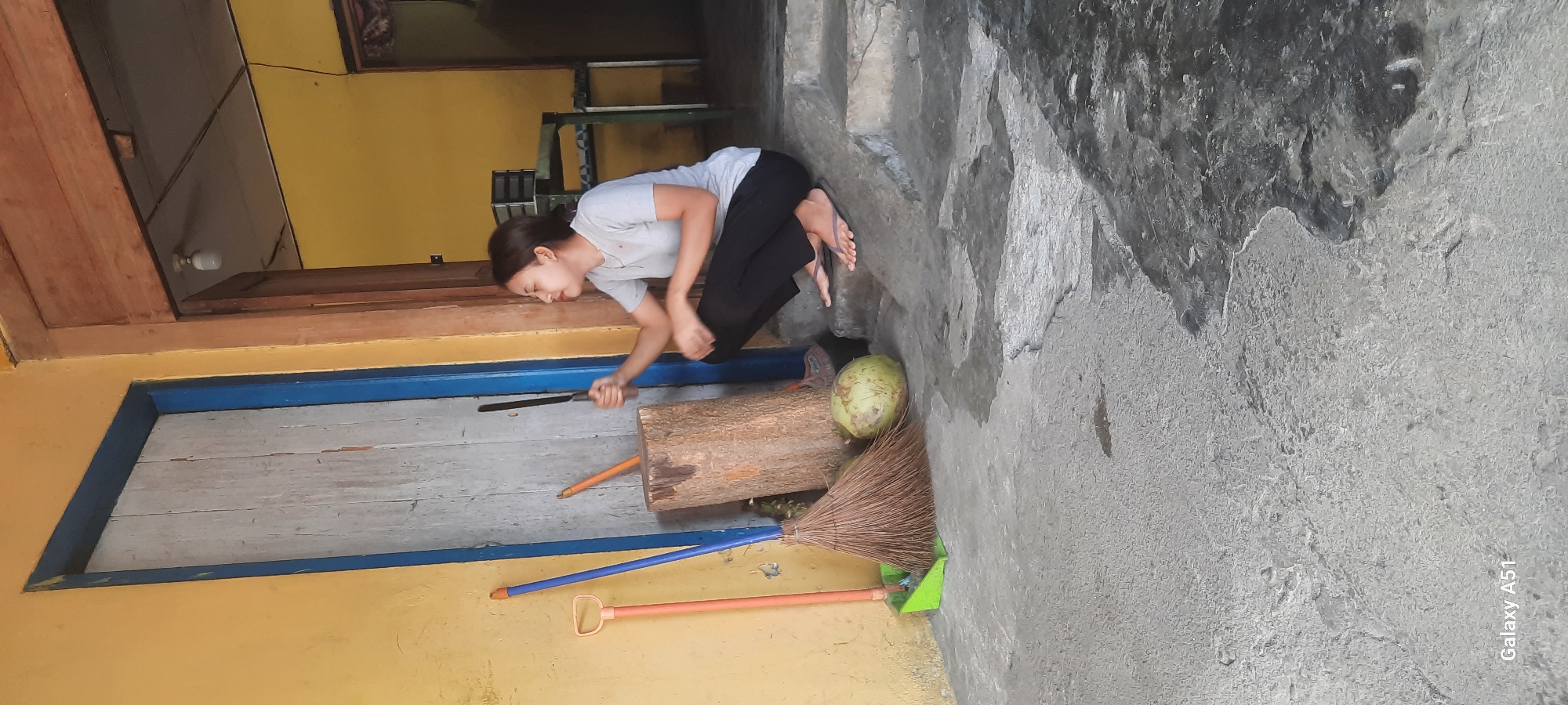 A young woman is squatting on concrete steps outside a building with yellow walls. She is wearing a light-colored t-shirt and dark pants. In her right hand, she holds a knife, and she appears to be holding it up to a large log. To her left, there is a coconut, a broom, and a dustpan. In the background, there is a wooden door with a blue frame, and beyond that, an open doorway reveals a glimpse of an interior space with yellow walls and a simple table and chairs. The scene appears to be set in Selopuro, Indonesia, likely in a residential or workshop area given the presence of tools and building materials.