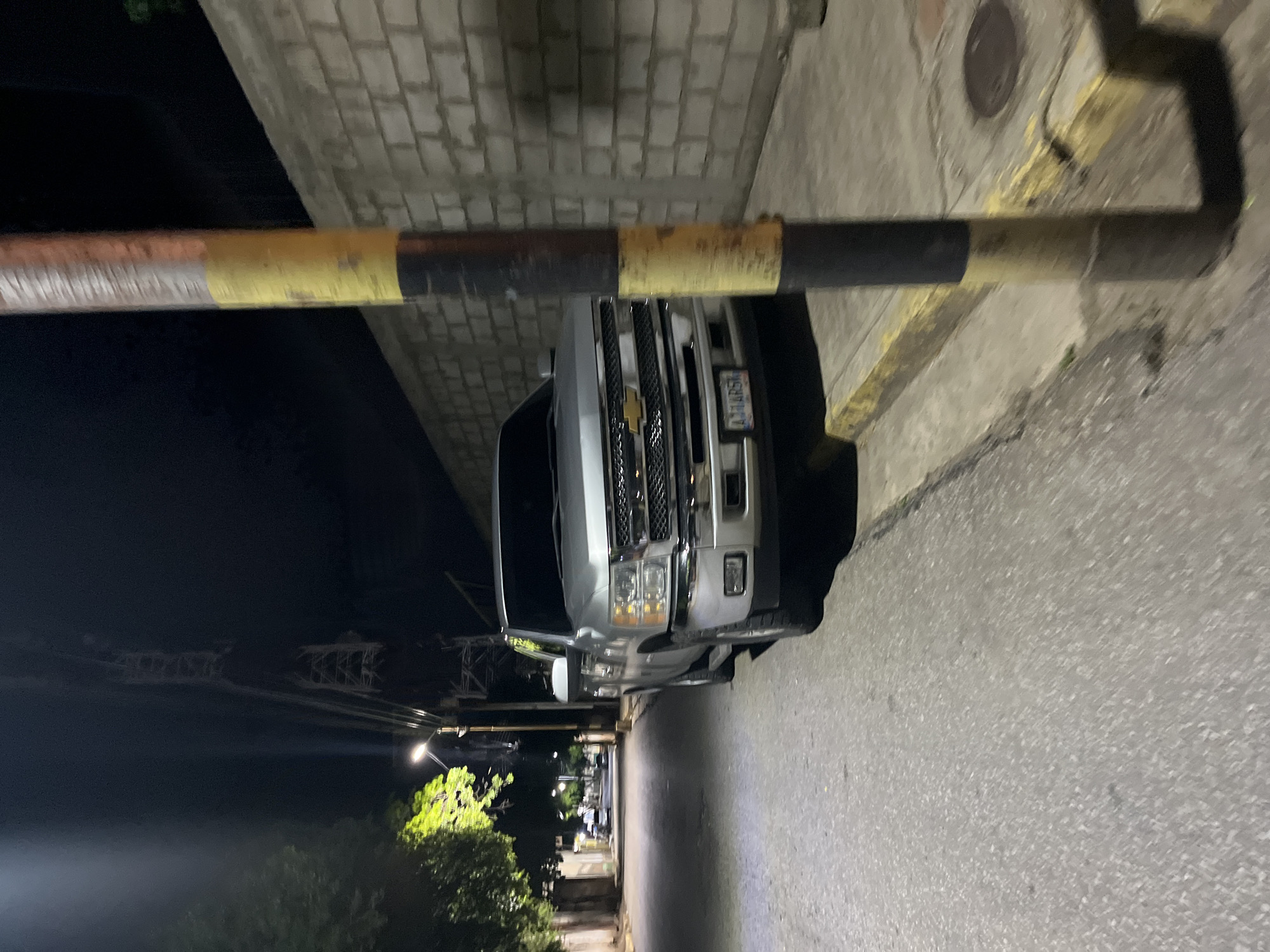 The image captures a nighttime scene on a street in Santa Ana, Venezuela. A silver Chevrolet Silverado pickup truck is parked on the right side of the road, facing forward. The truck's headlights are off, and its license plate is partially visible. A striped pole, with black and yellow bands, stands on the sidewalk next to the truck. Behind the truck and pole, a wall constructed of concrete blocks runs along the property line. The street is paved, and a sidewalk is visible. Trees with illuminated foliage are present on the left side of the street, suggesting the presence of streetlights or architectural lighting. In the background, streetlights illuminate the road, and a large industrial structure, possibly a crane or tower, is visible against the dark night sky. The overall atmosphere is quiet and still.