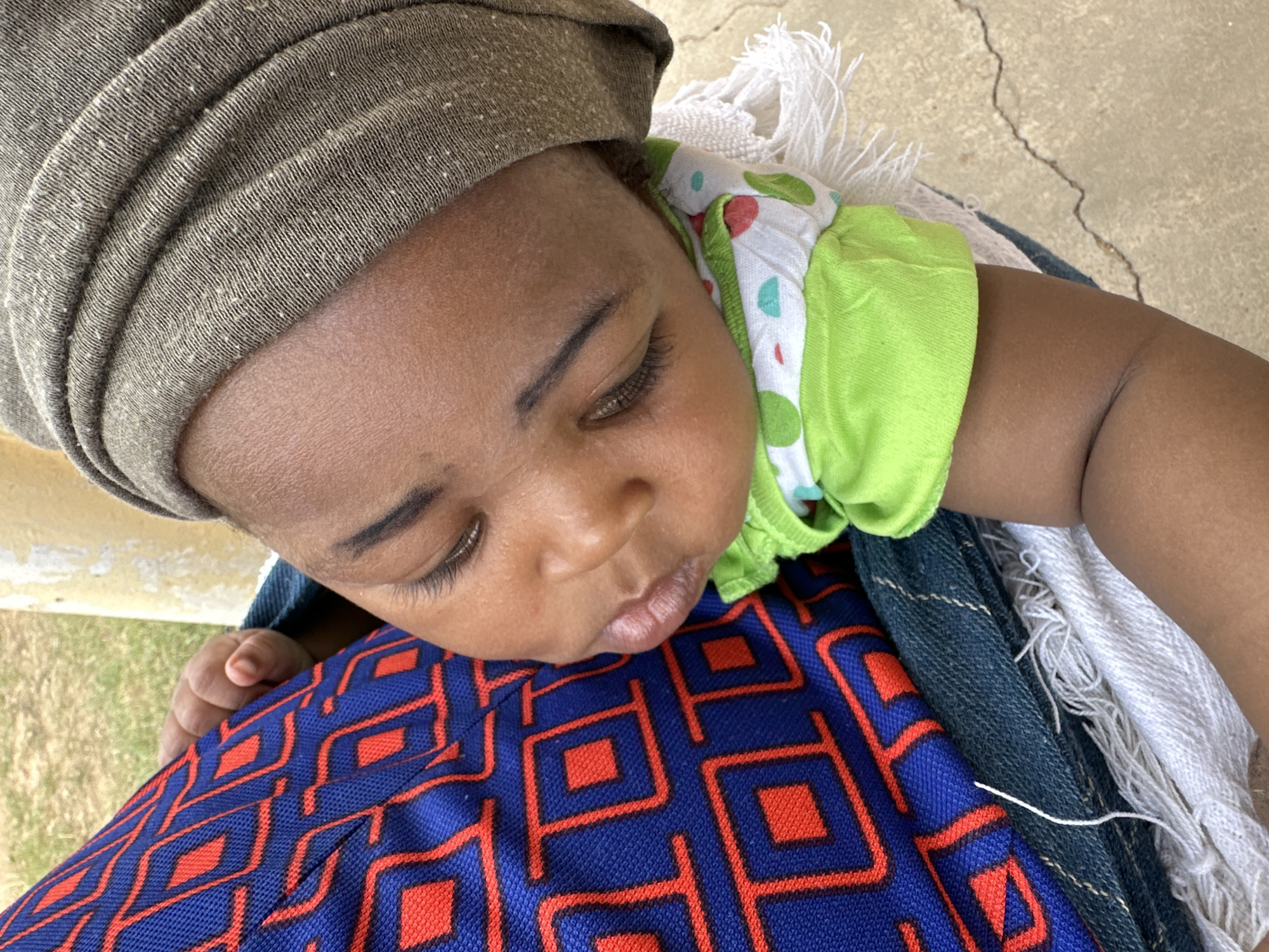 The image presents a high-angle, close-up view of a baby resting peacefully against an adult. The baby, with dark skin, has their eyes closed, showcasing long, dark eyelashes, and their lips are slightly parted. They are wearing a light green top adorned with a pattern of white, red, and light blue dots. A greyish-brown head wrap or turban covers the baby's head.
The baby is leaning against what appears to be an adult's chest or lap. The adult's clothing features a vibrant blue fabric with a prominent repeating red geometric pattern, suggesting a traditional garment. Below this, a dark blue, possibly denim, fabric is visible, alongside a white or off-white textured fabric with loose threads or fringes. A small, closed fist of the baby can be seen resting on the blue patterned fabric.
The background includes a light-colored, textured surface, resembling concrete or pavement, with a distinct crack. Patches of green grass or vegetation are visible in the lower left, indicating an outdoor or semi-outdoor setting. The bright, natural lighting suggests the photograph was taken during the daytime. The scene conveys a sense of calm and comfort for the sleeping baby. No text is visible in the image. The clothing styles and outdoor environment are consistent with the provided location context of Jos, Nigeria.