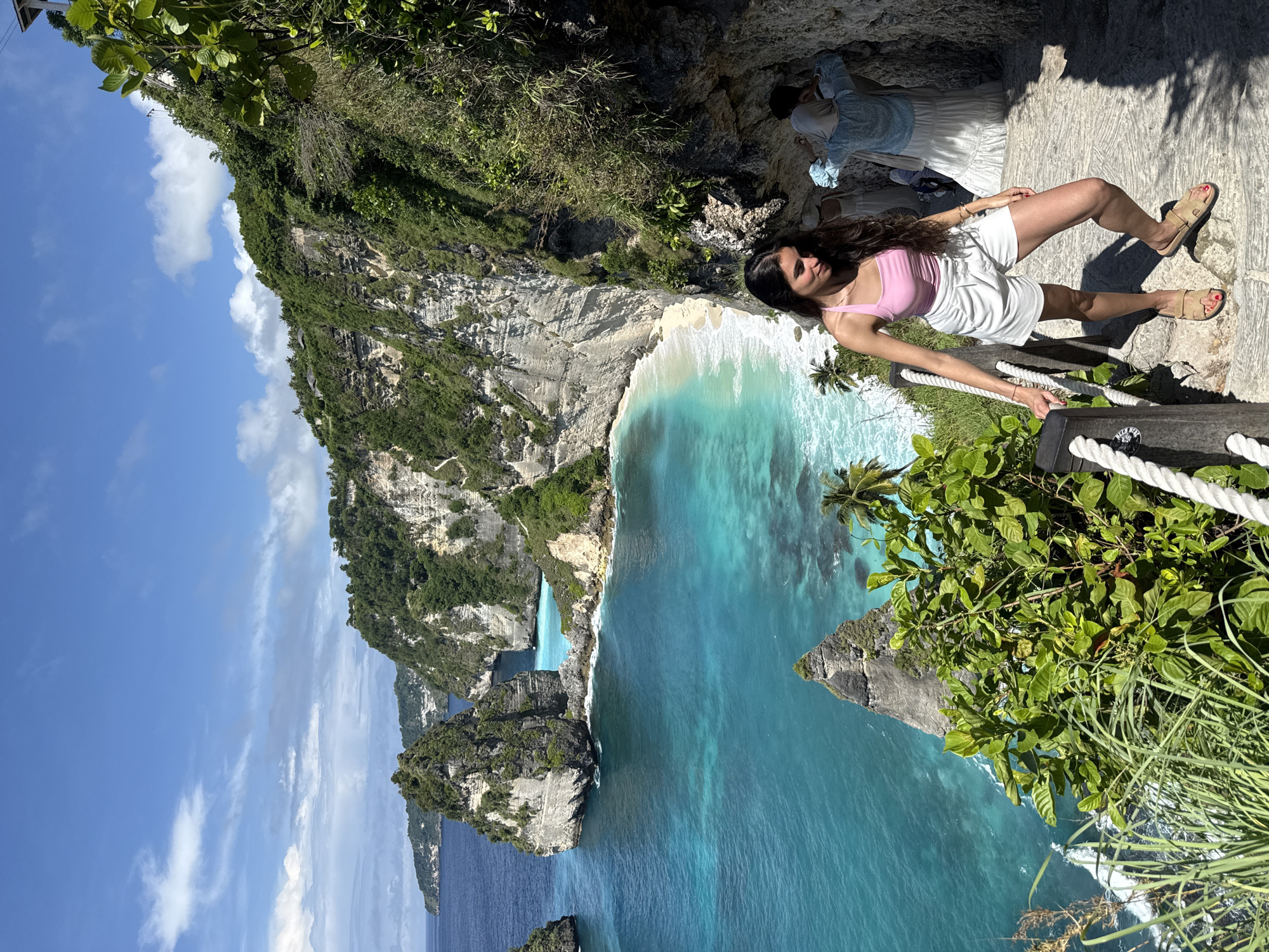 A woman lies on a stone walkway in Pejukutan, Indonesia. She wears a pink tank top, white shorts, and sandals. She leans against a white rope railing. The ocean stretches out below. A cliff face rises at the left. Green vegetation surrounds the scene. The sky is blue with white clouds. Two other people can be seen on the stone walkway in the background.