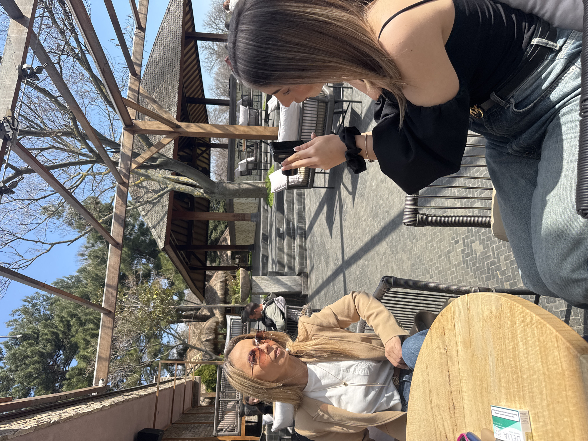 Two women are seated at a round wooden table in an outdoor setting, with one woman holding a phone and the other looking at the camera. The woman with the phone is wearing a black top and jeans. The other woman is wearing a white shirt, jeans and a beige blazer and pink tinted sunglasses. There is a pack of cigarettes on the table. The location appears to be an outdoor restaurant or café in Varna, Bulgaria, characterized by tiered seating, thatched roof structures, wooden beams, and trees. There are other people seated in the background. The photo is taken during the day with a bright sky.