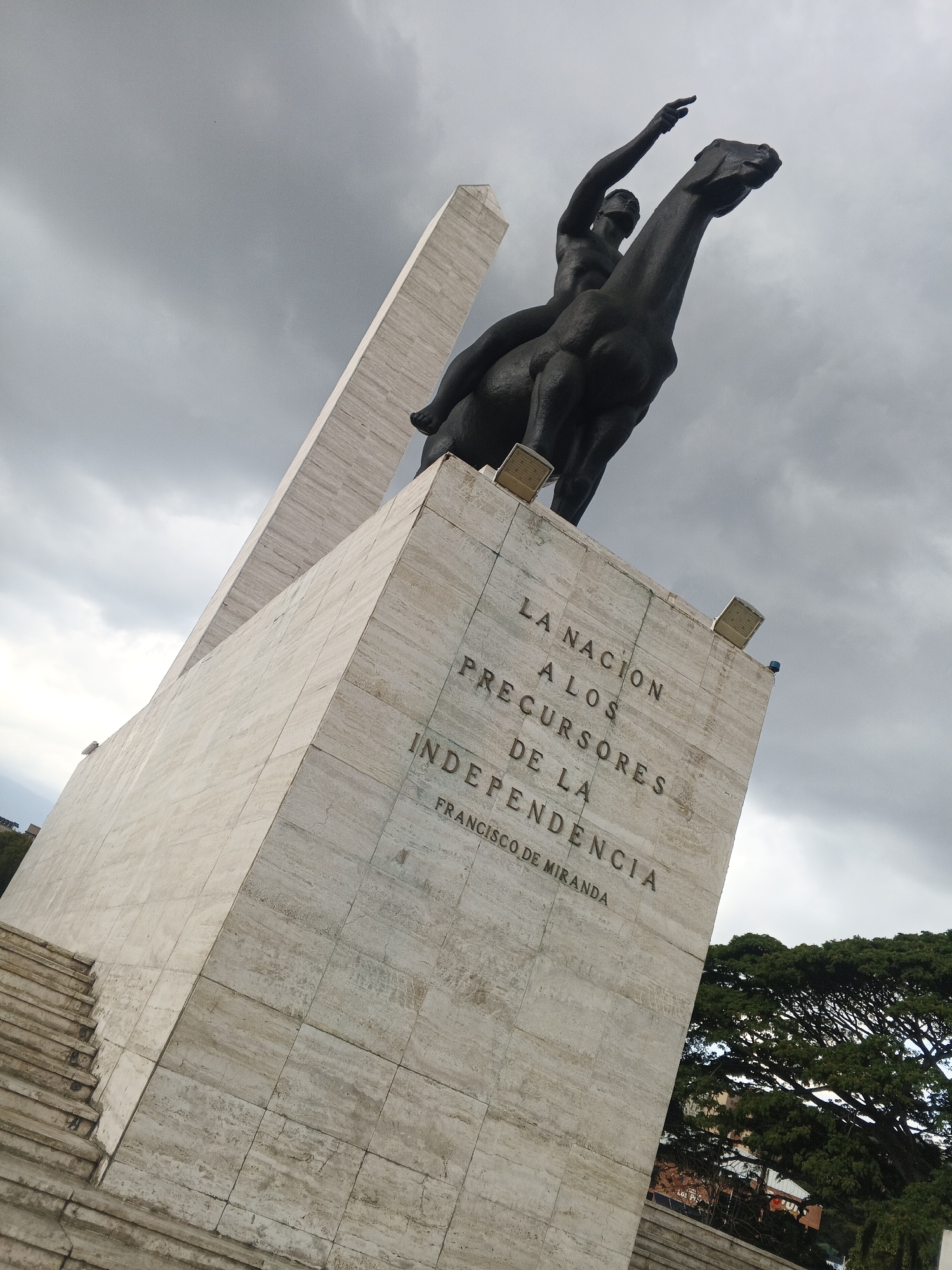 A low-angle shot captures a tall, light-colored stone monument against a cloudy sky. The monument features a bronze equestrian statue of a rider on horseback. The rider is shirtless, with one arm raised. Inscribed on the monument's stone are the words "LA NACION A LOS PRECURSORES DE LA INDEPENDENCIA. FRANCISCO DE MIRANDA." A staircase is visible on the left side of the monument, and a large tree with dense foliage is visible on the right. The statue is located in the city of Unknown, Unknown.