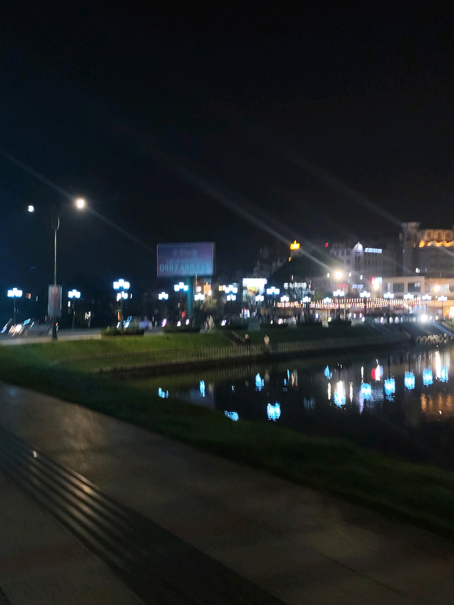The image depicts a nighttime scene along a waterfront in Da Lat, Vietnam. A paved walkway with textured strips runs diagonally across the foreground, bordered by a grassy area. Across the water, the city skyline is illuminated by numerous lights from buildings and street lamps, creating reflections on the water's surface. A billboard with Vietnamese text and a phone number is visible on the left side of the frame. Several streetlights with multiple bulbs are positioned along the walkway, casting light onto the scene. The overall impression is a lively urban environment at night, with the water acting as a reflective canvas for the city's glow.