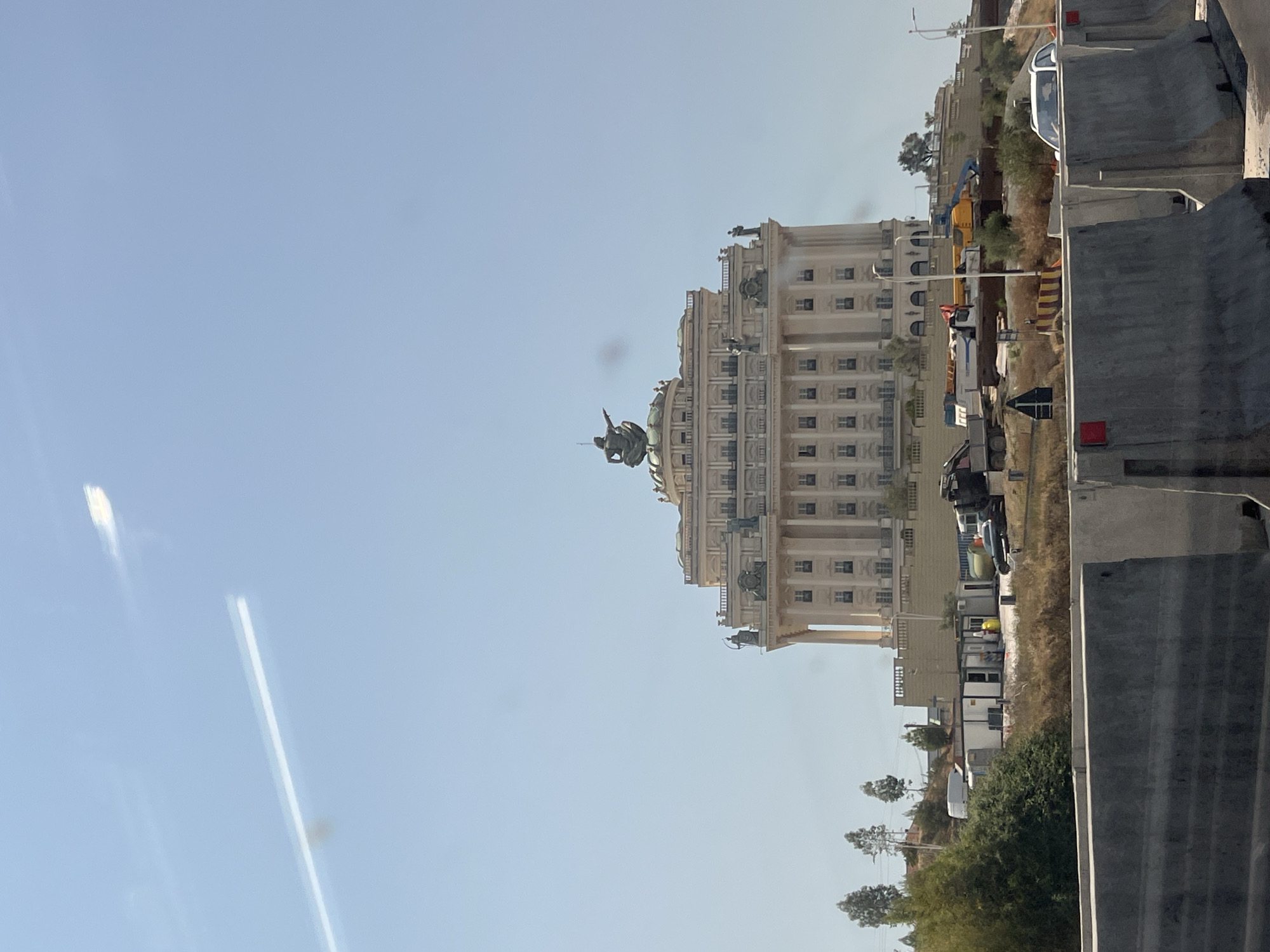 A large, ornate, multi-story building with classical architectural elements, including columns and decorative statues, dominates the upper half of the frame. A bronze statue of a seated figure pointing is positioned on the building's dome. The building is situated on a hillside overlooking a construction area. In the foreground, a row of grey concrete barriers is visible, suggesting a road or highway. The environment is arid, with dry grass and sparse trees. Construction vehicles, including a truck and what appears to be a crane, are present in the middle ground. The sky is clear and light blue, with a faint light streak visible in the upper left quadrant. The building's design is reminiscent of grand public structures. The overall scene depicts a blend of historical architecture and ongoing development in Kashar, Albania.