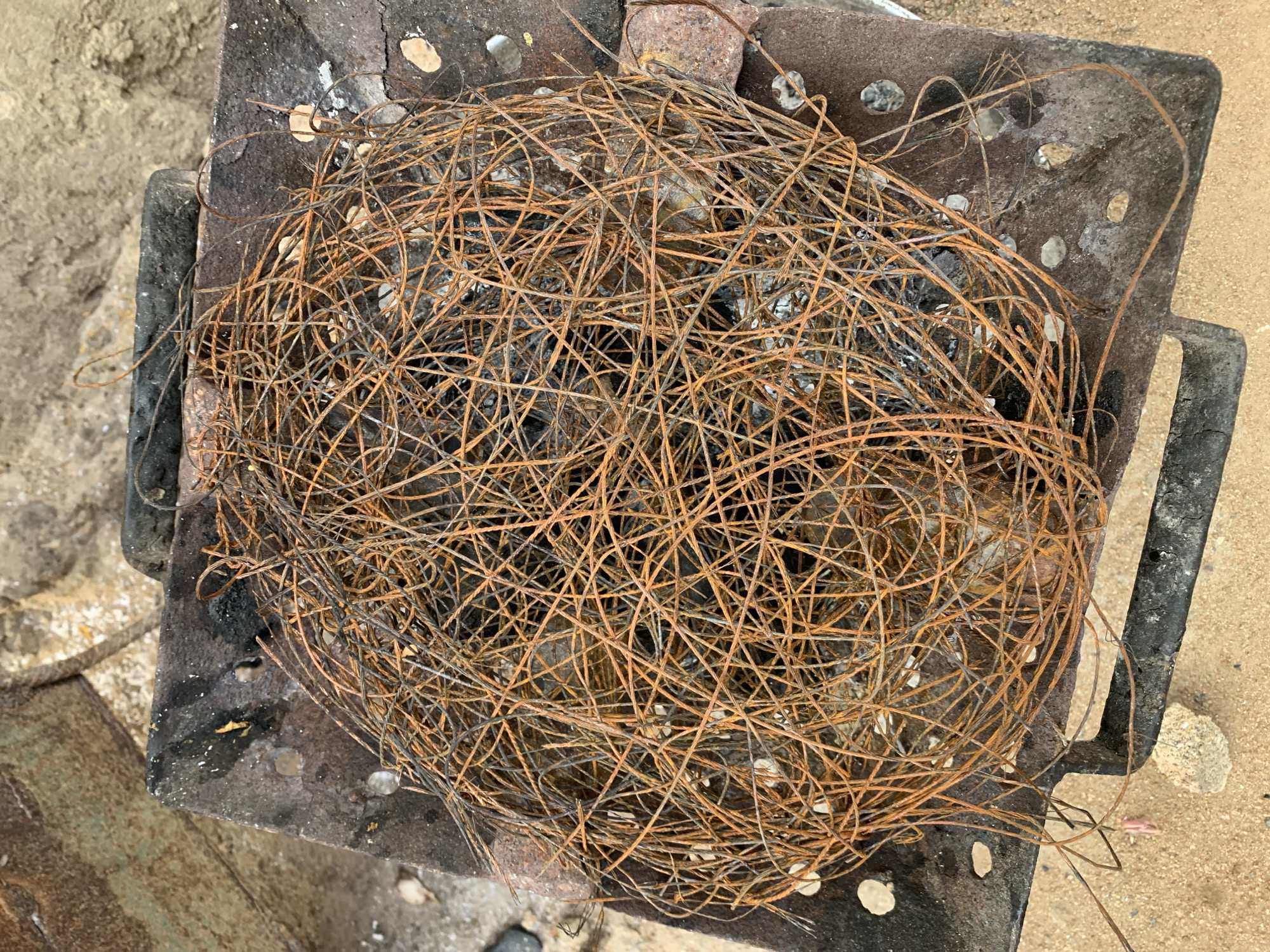 A large, tangled mass of thin, oxidized metal wires, predominantly orange-brown with some darker sections, occupies the central area of the image. This wire cluster rests within a dark, rectangular metal structure, possibly an improvised brazier or grate, which exhibits multiple irregular perforations, charring, and areas of discoloration. The structure is situated on a light brown, sandy ground. In the upper left background, a corroded, greenish-brown metal surface, indicative of a larger object or structure, is partially visible. Additional dark, rectangular elements, possibly support blocks or handles, are present along the perimeter of the primary metal structure. Small, light-colored fragments, consistent with ash or pebbles, are scattered across the surface of the metal structure and the surrounding sand. No human figures or direct actions are depicted. The scene is located in New Stadium, Nigeria.